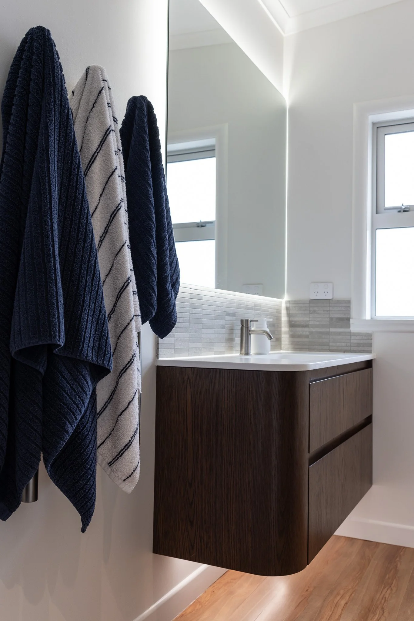 A modern bathroom with a floating dark wood vanity, a white countertop, a single faucet, and a mirror reflecting a window. Three towels hang on a rail next to the mirror, with two dark blue and one white with black stripes. The room has a sloped ceil