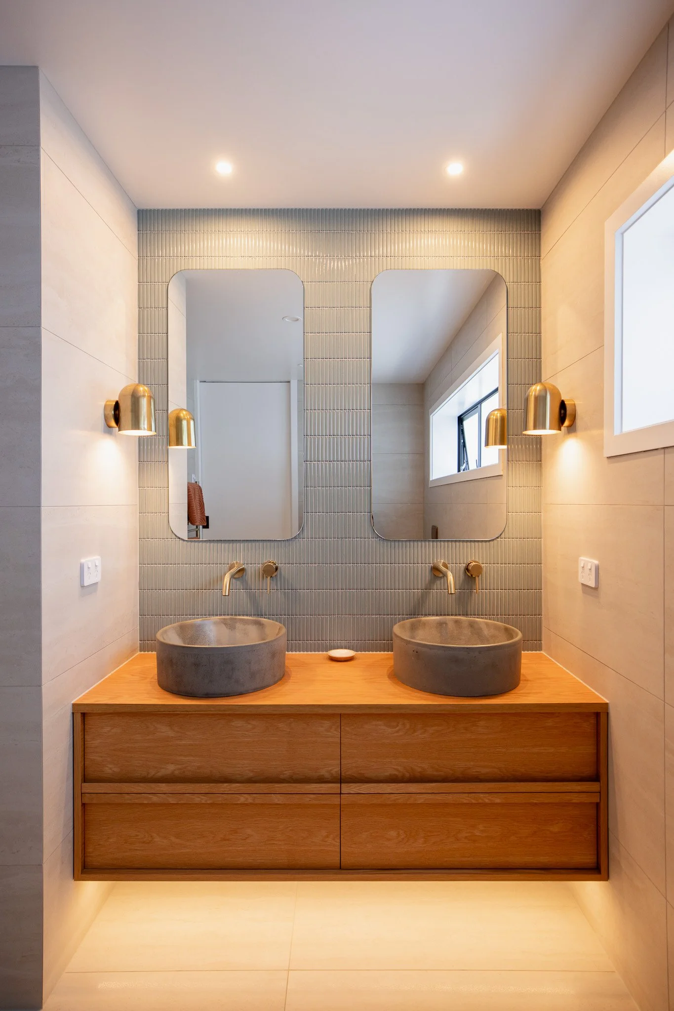 Modern bathroom vanity with two concrete vessel sinks, large mirrors, and mounted gold-colored wall sconces, with a window on the right side.