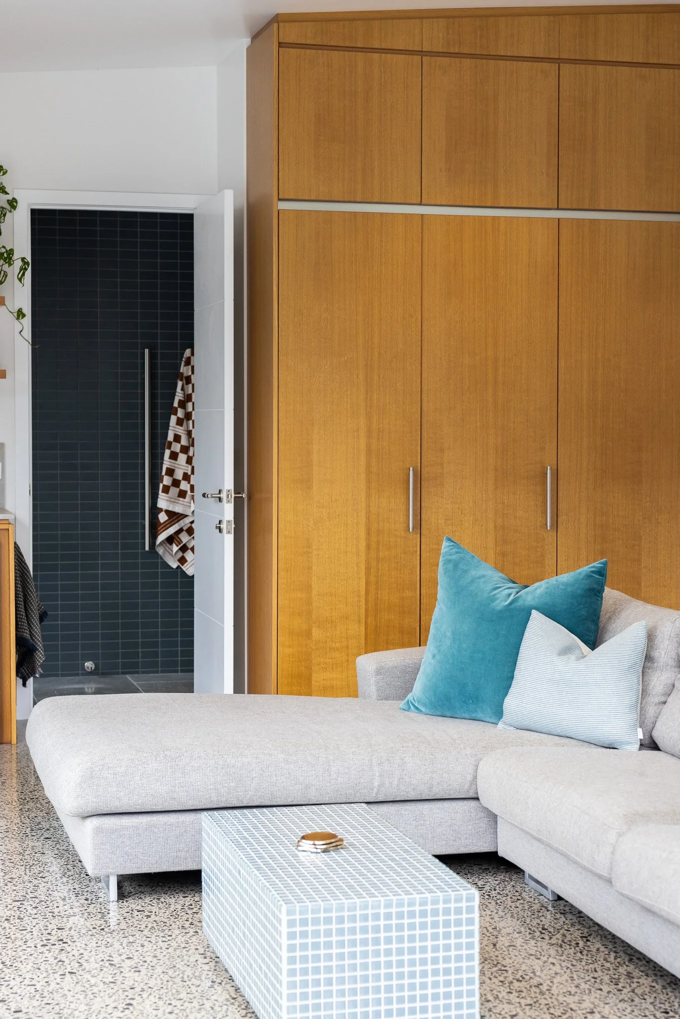 Living room with beige sectional sofa, blue and white pillows, white tiled coffee table, wooden closet, and open door revealing a dark bathroom with black tiles and a towel hanging.