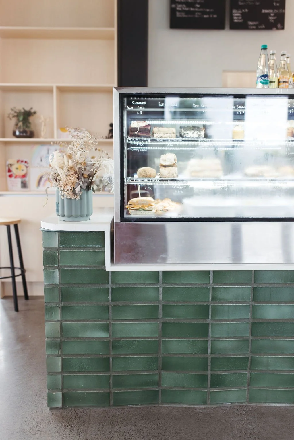 Display case with assorted desserts such as cakes and pastries, with a vase of dried flowers beside it, in a modern cafe setting.
