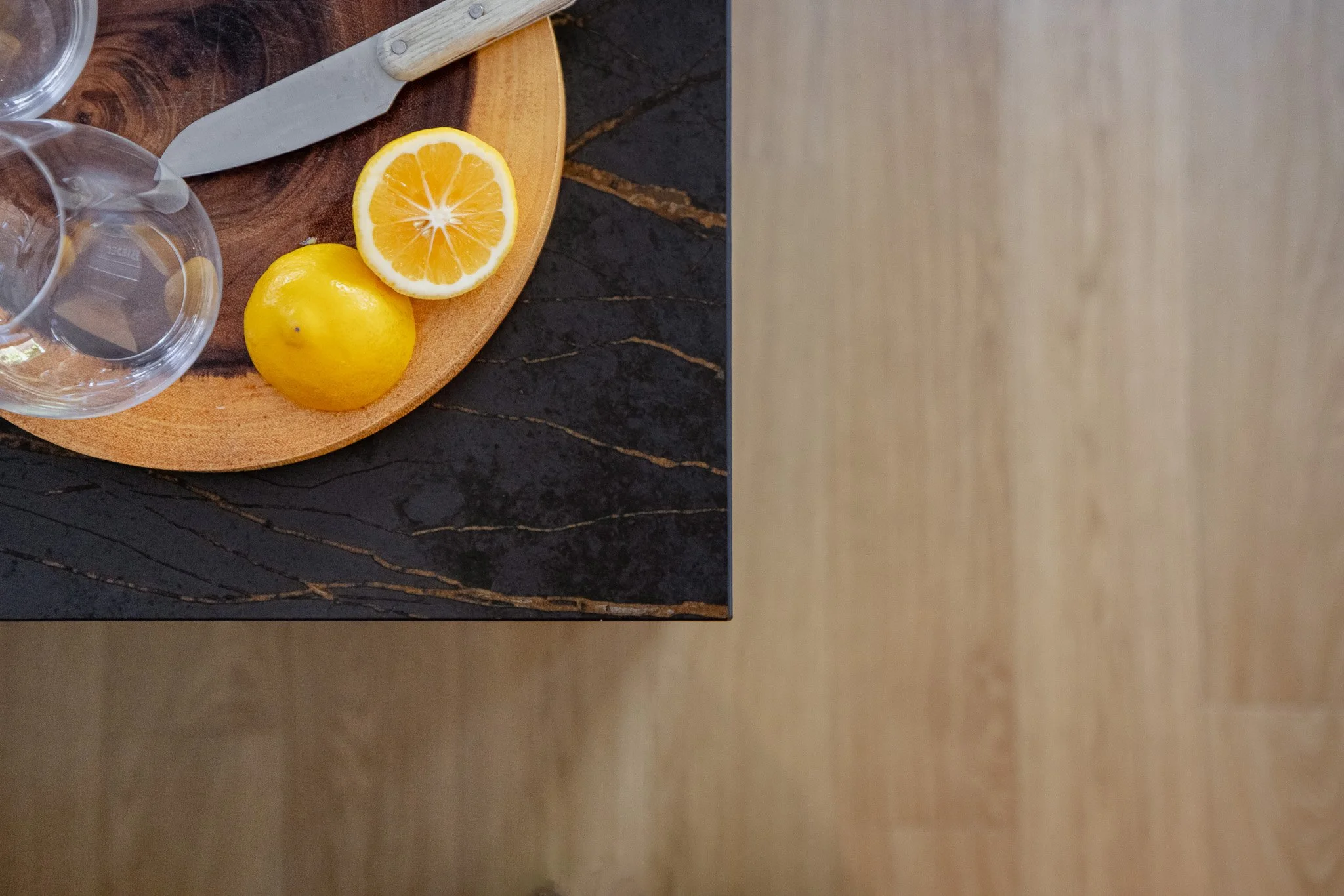 A lemon and a lemon half on a round wooden cutting board next to a glass of water and a knife, on a dark marble countertop.
