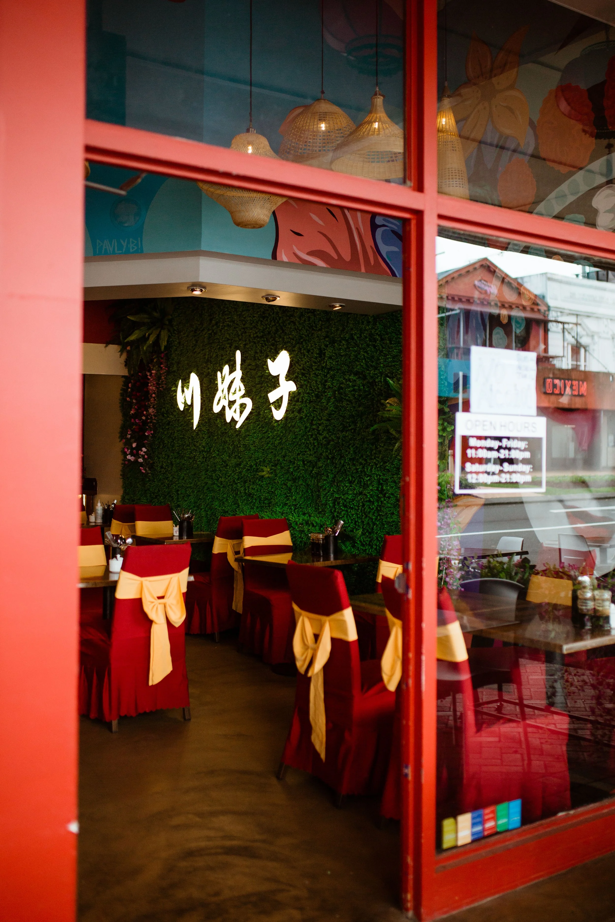 Inside a restaurant with red chairs adorned with gold sashes, a green wall with white Asian characters, and hanging woven lamps, viewed through a red door window.