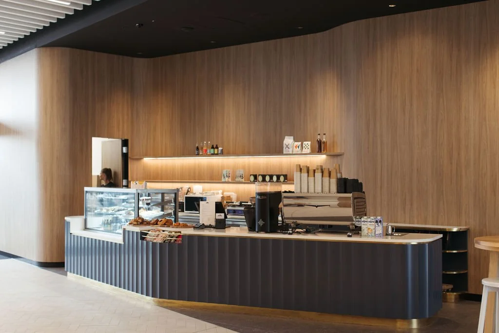 A modern cafe counter with a glass display of baked goods, coffee machines, and coffee cups in a stylish setting with wooden walls and black accents.