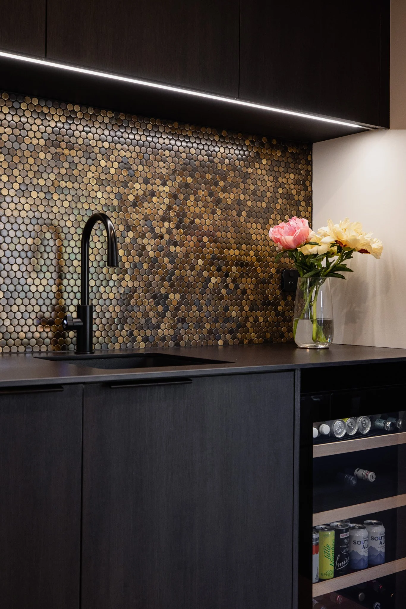 Modern black kitchen with a gold mosaic tile backsplash, black sink and faucet, and a glass vase with pink and cream peonies on the counter.