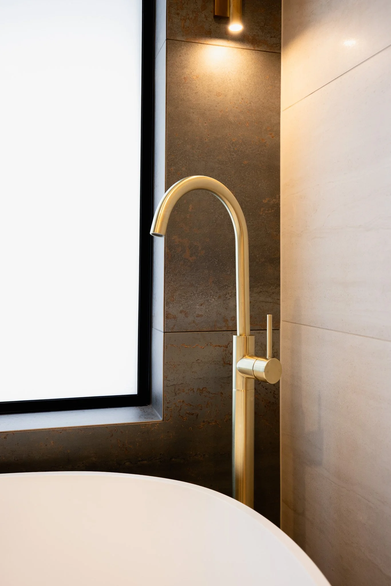 Close-up of a gold-colored modern faucet next to a bathtub with a frosted window in the background.