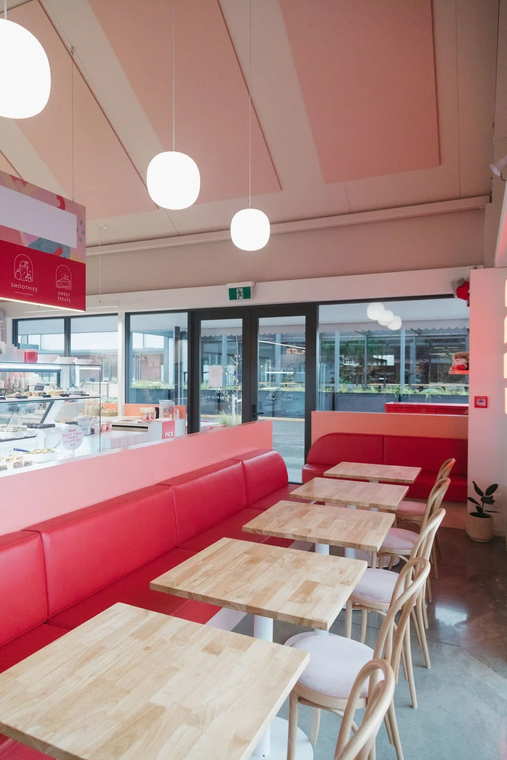 Interior of a modern cafe with red seating, light wooden tables, and chairs. The ceiling has pink acoustic panels and white pendant lights. Large windows provide outdoor views, and there's a display fridge with food visible.