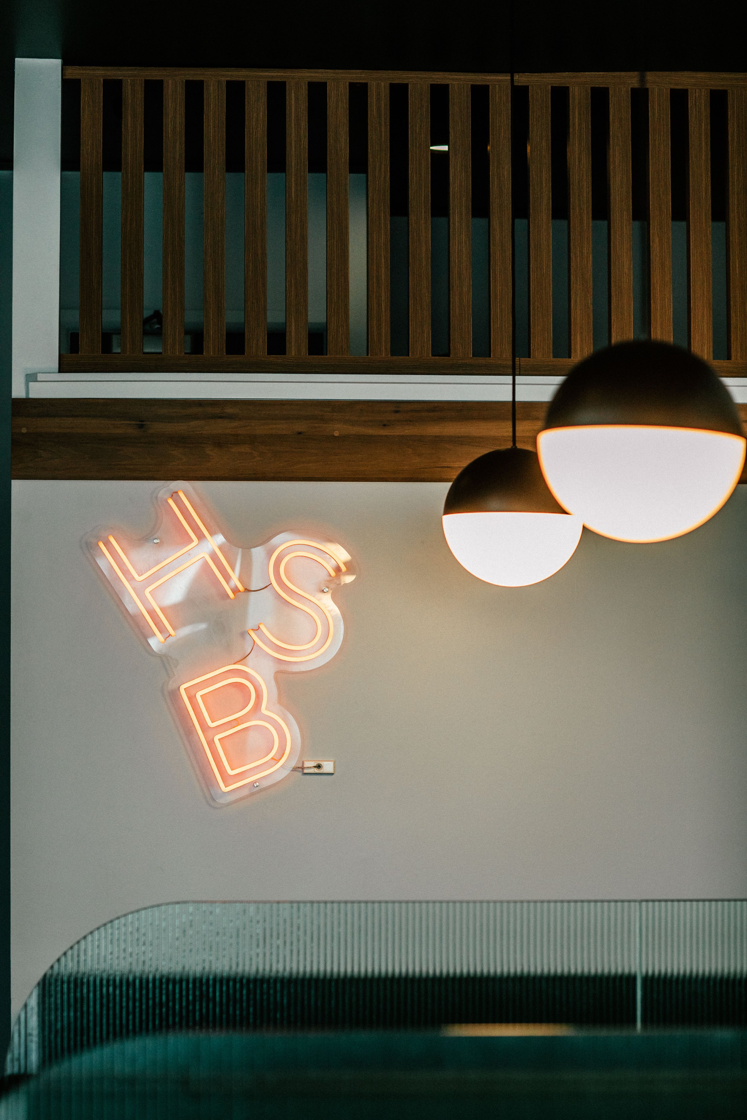 Neon sign with the words 'HBS B' on a white wall, two hanging spherical pendant lights above, wooden railing in the background.