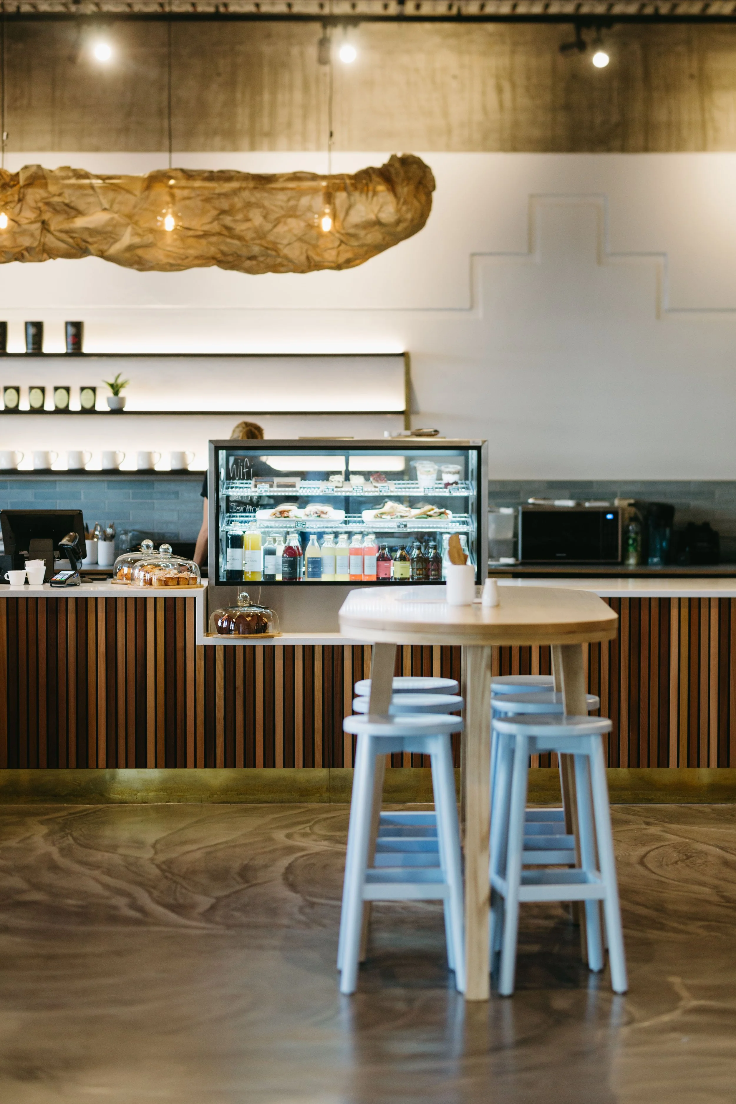 Interior of a modern cafe with a coffee counter, white stools around a small round table, and warm lighting.
