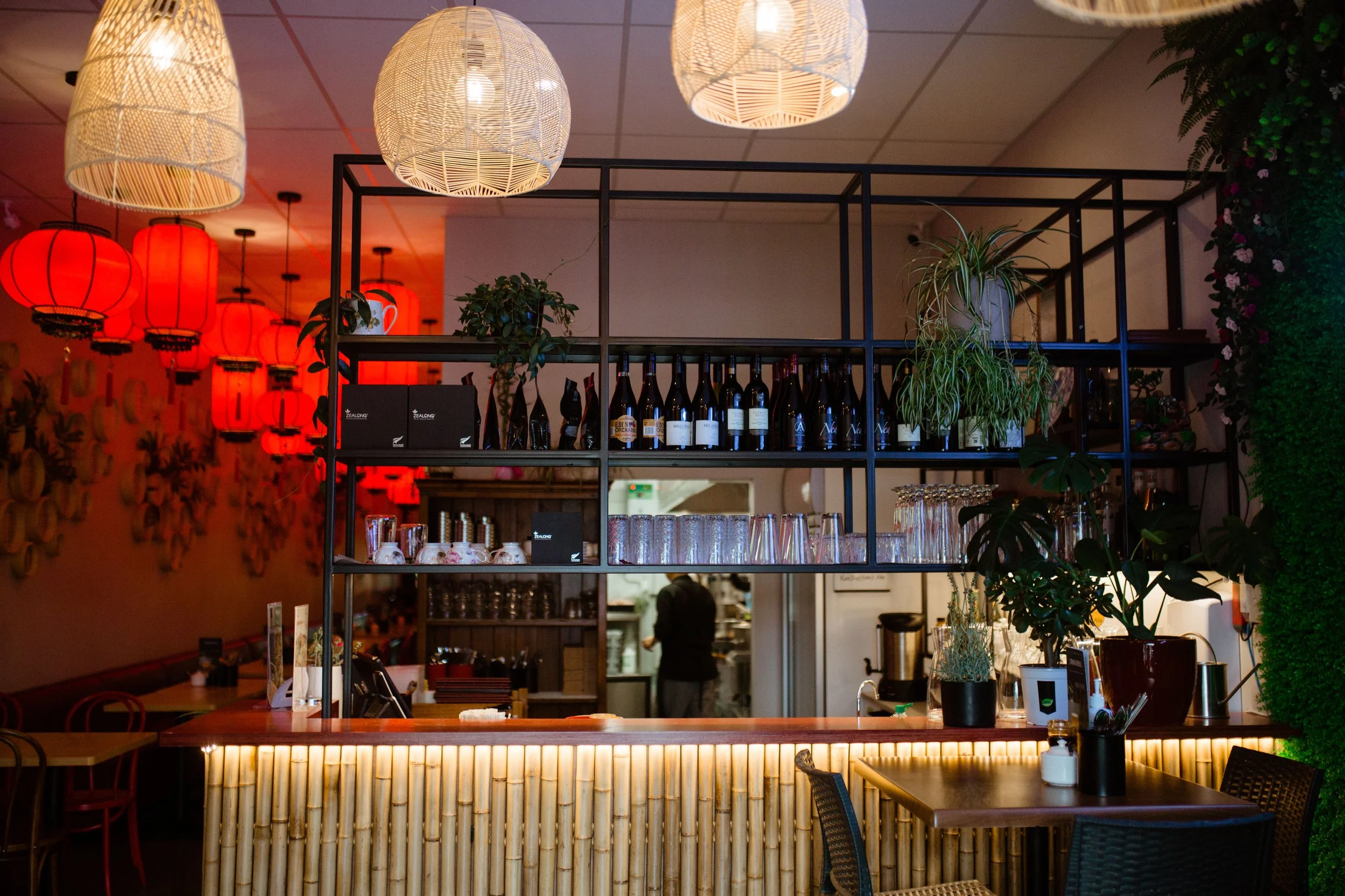 A restaurant or bar interior with a bamboo-covered counter, hanging woven and red lantern lights, potted plants, and a shelving unit with bottles, glasses, and decor. A person works in the kitchen in the background.