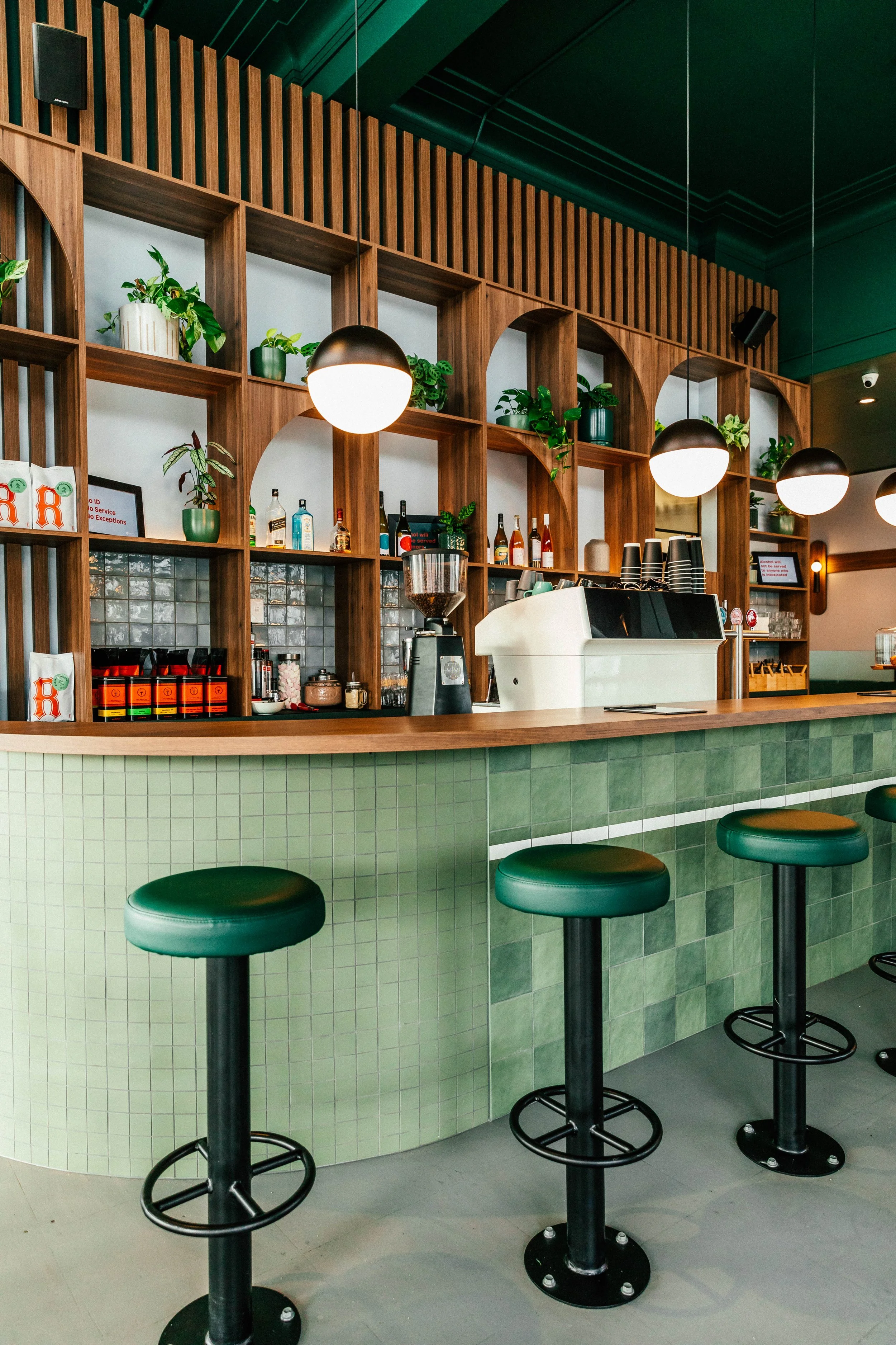 Interior of a modern coffee shop with green bar stools, green tiled bar front, and wooden shelves with plants and bottles behind the counter.
