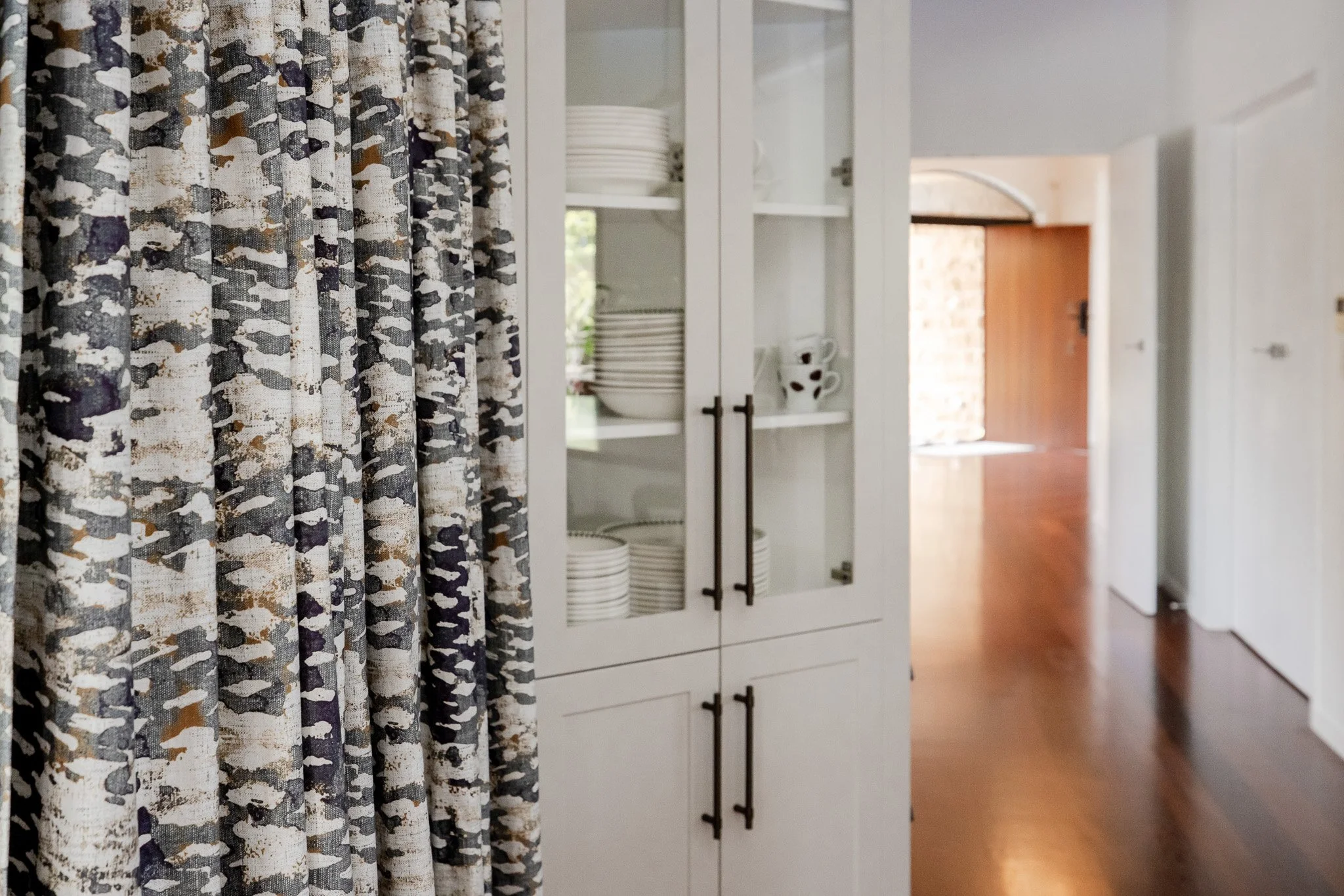 A white kitchen cabinet with glass doors, filled with stacked white dishes and cups, near a hallway with wooden flooring and a window with patterned curtains.