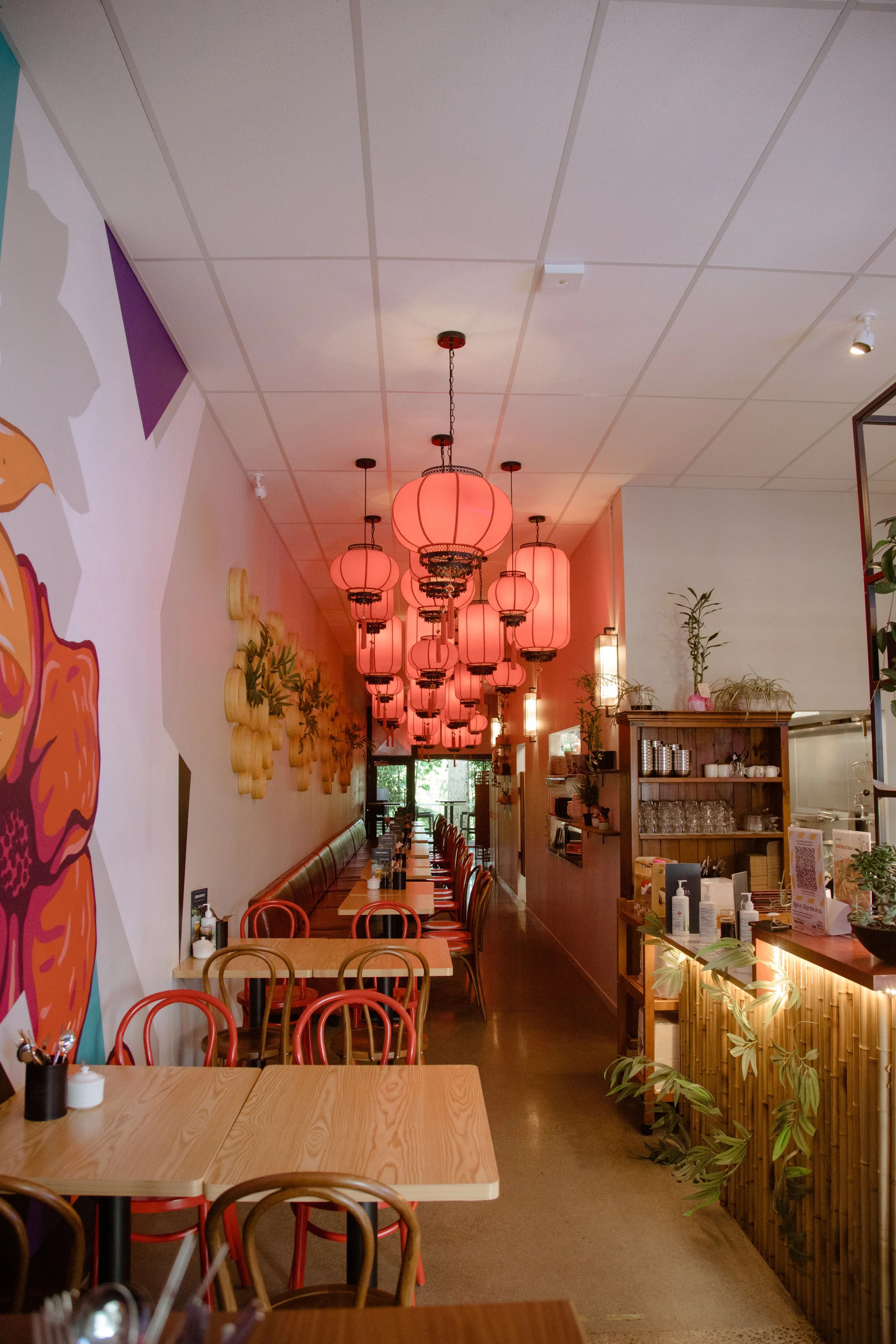 Interior of a restaurant with wooden tables and chairs, pink lantern ceiling lights, and wall decorations with plants and art.