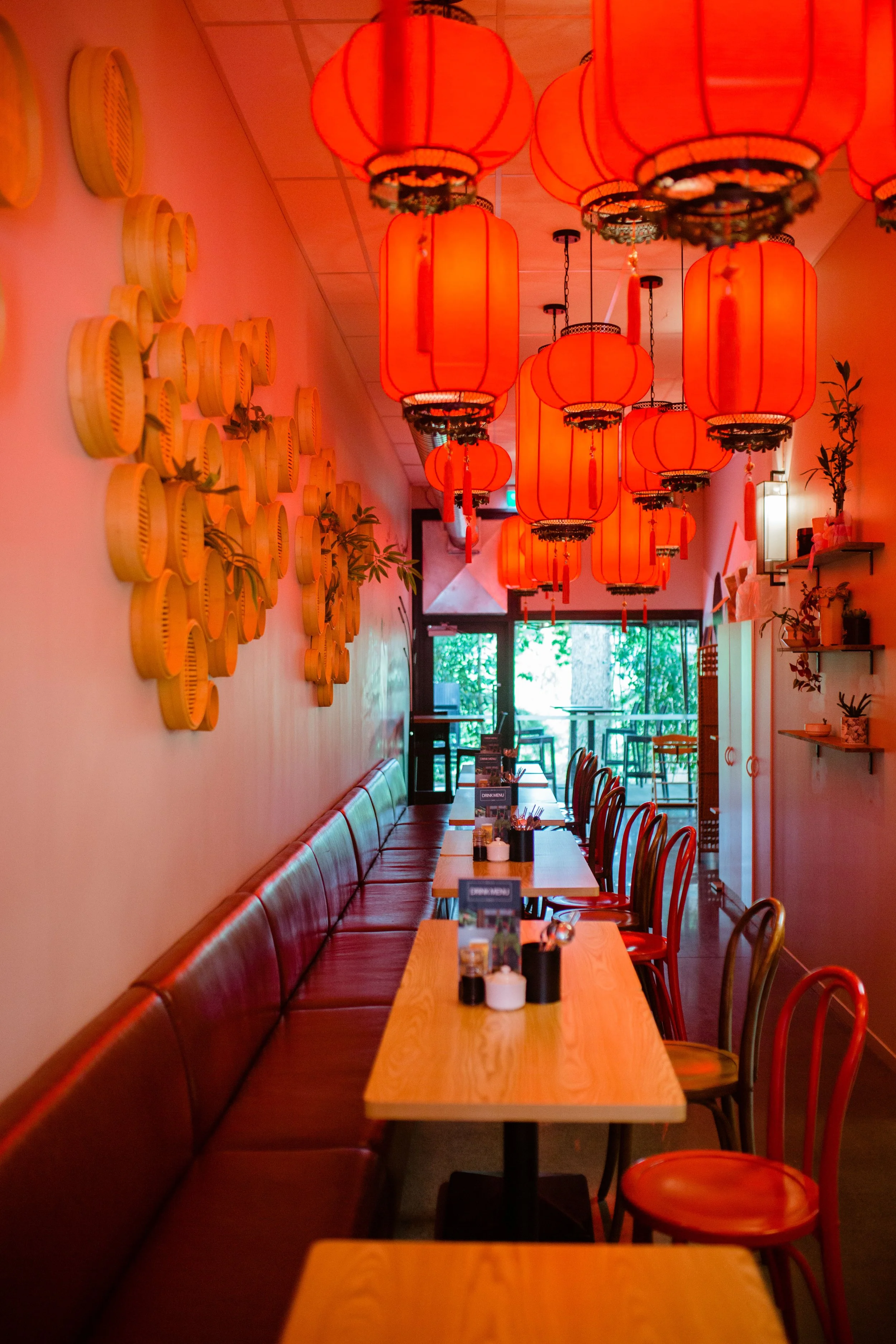 Interior of a restaurant decorated with red Chinese lanterns hanging from the ceiling, a long wooden table with chairs, and wall decorations including a yellow wall art piece and shelves with plants.