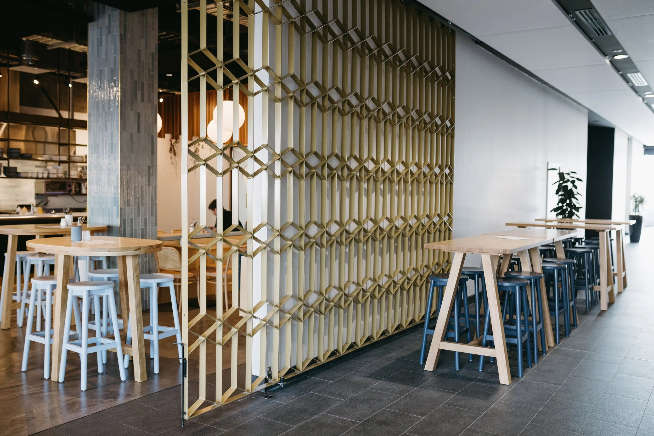 Modern restaurant interior with wooden tables and stools, a decorative wooden partition, indoor plants, and large windows letting in natural light.
