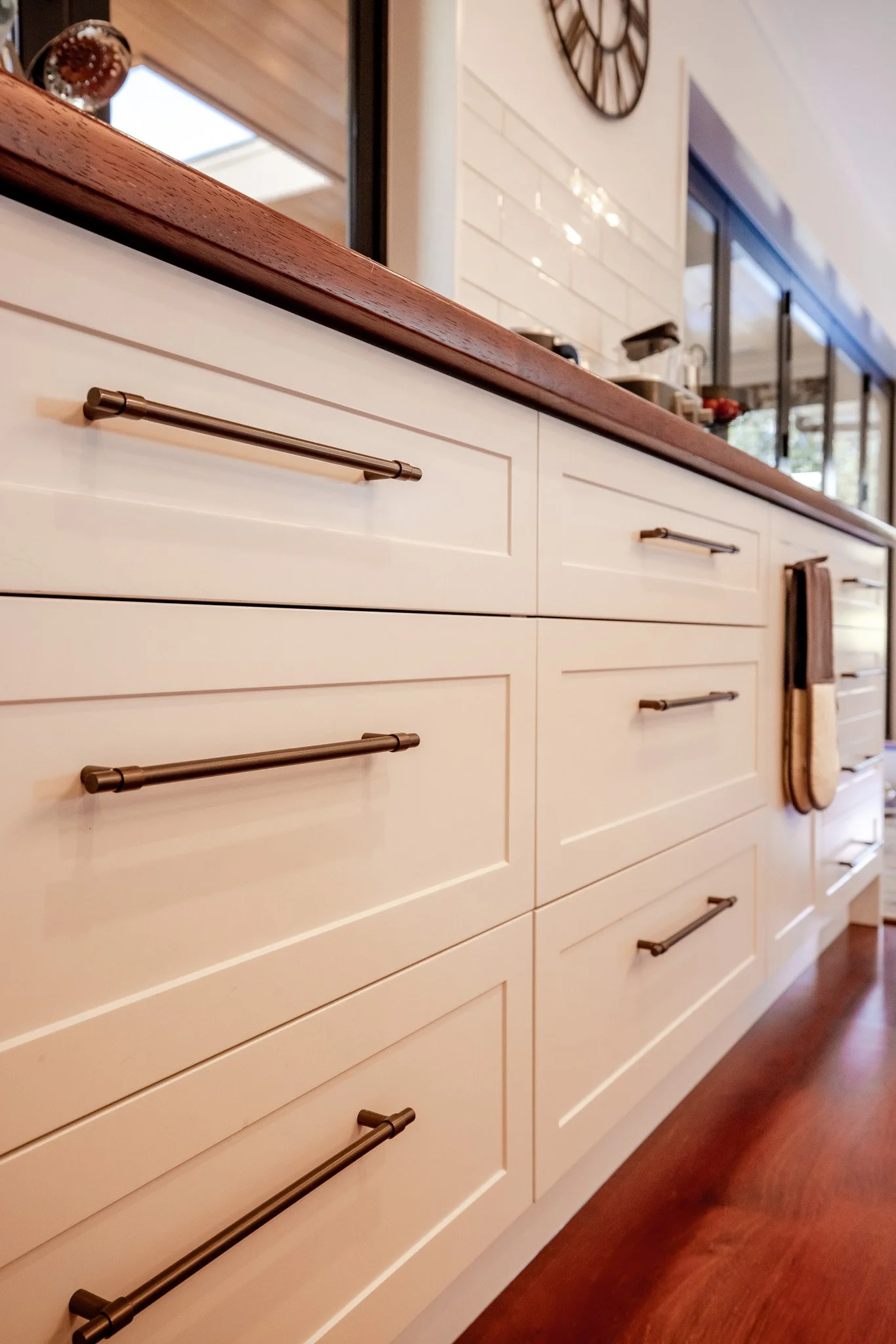 Close-up of white kitchen cabinets with black metal handles and a wooden countertop, with a mirror and wall clock visible in the background.