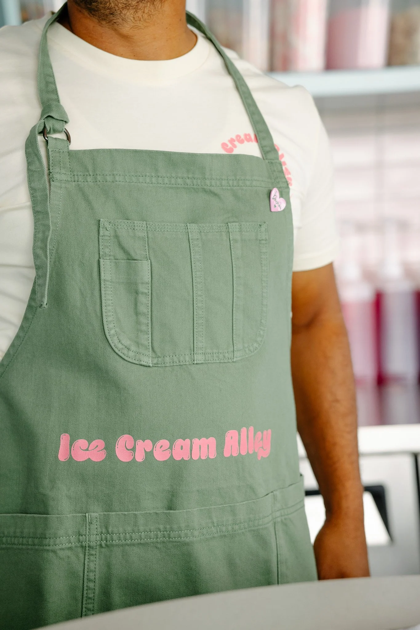 Close-up of a person wearing a green apron with 'Ice Cream Alley' written in pink, over a white t-shirt with red print. Part of the background shows shelves with pink containers.