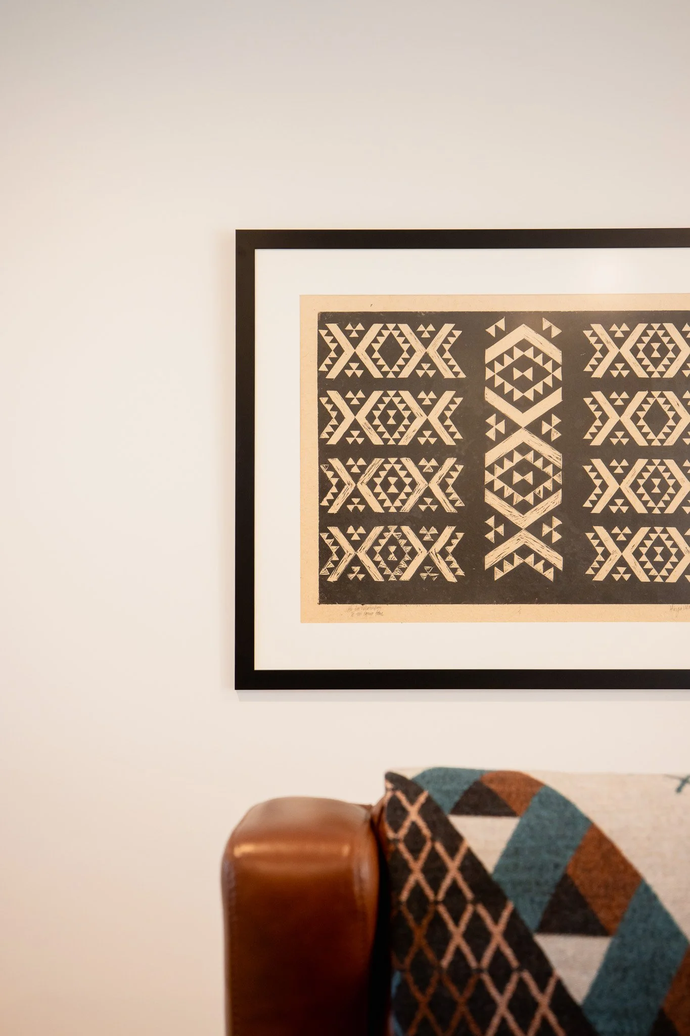 Part of a leather-cushioned chair with a geometric patterned fabric cover, against a white wall with a framed black-and-white geometric artwork.