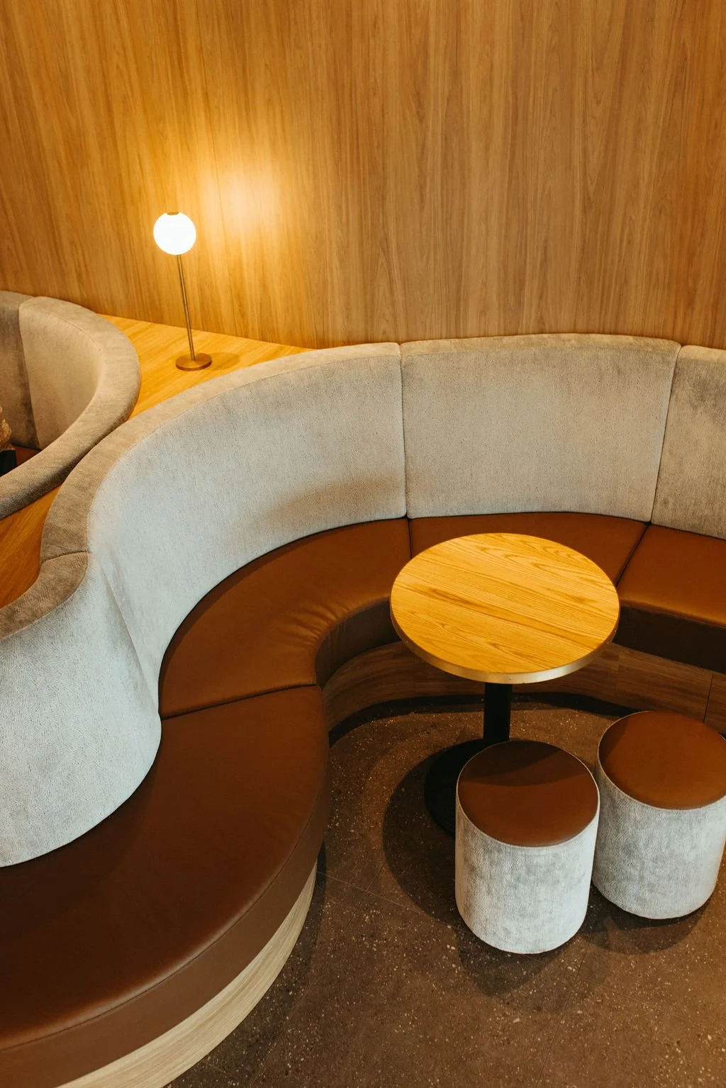 Curved beige and brown upholstered booth seating with a small circular wooden table and matching round stools, with a wooden wall and a small modern lamp in the background.