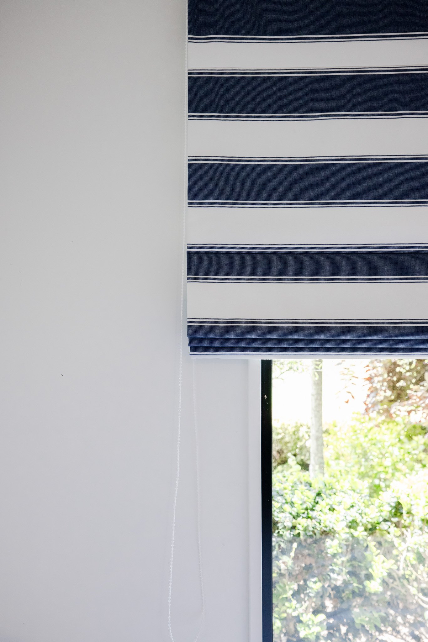 A window with a white wall and a partially drawn blue and white striped Roman shade or curtain.