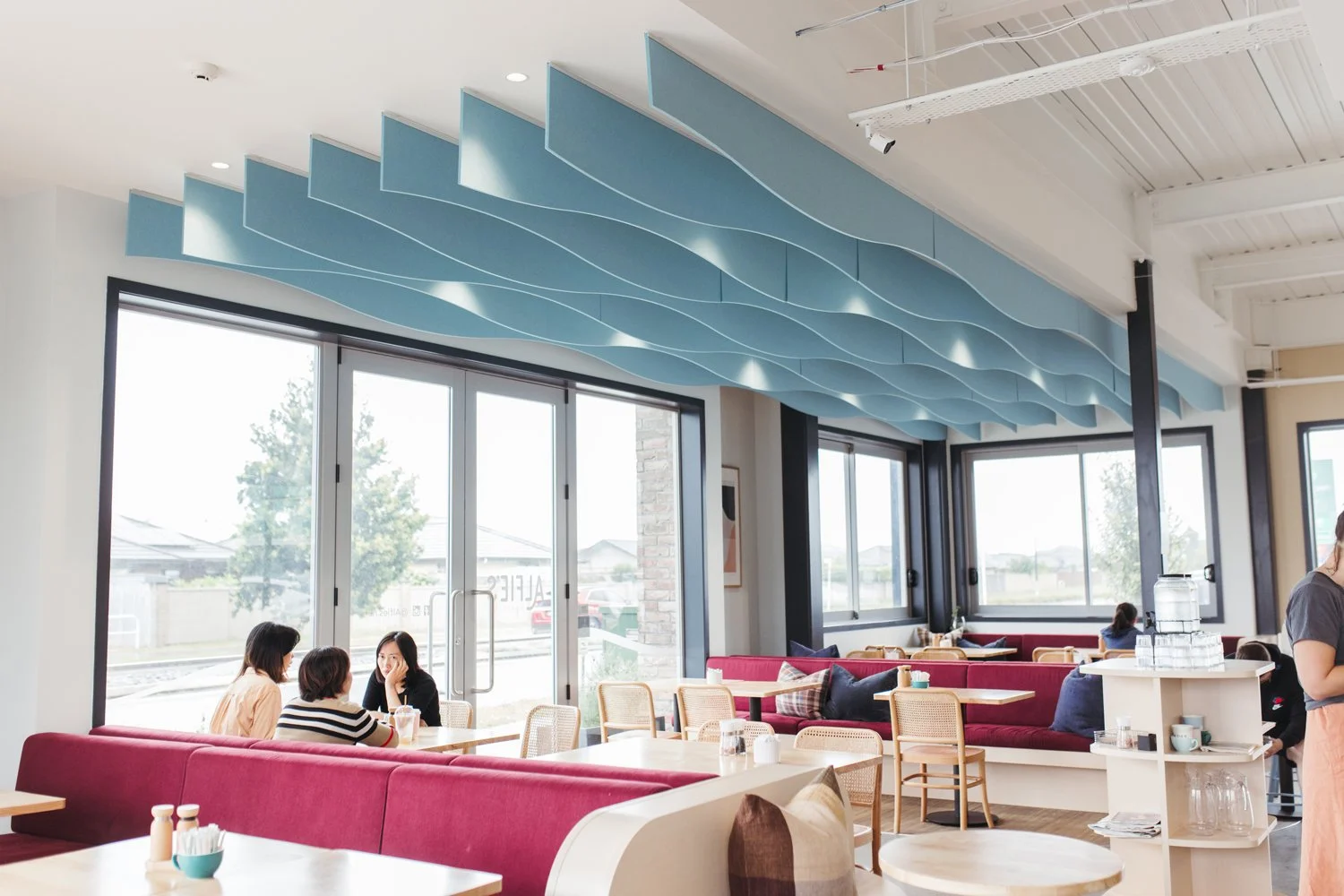 Interior of a modern cafe with pink and beige seating, wooden tables, large windows, and a wavy blue ceiling installation.