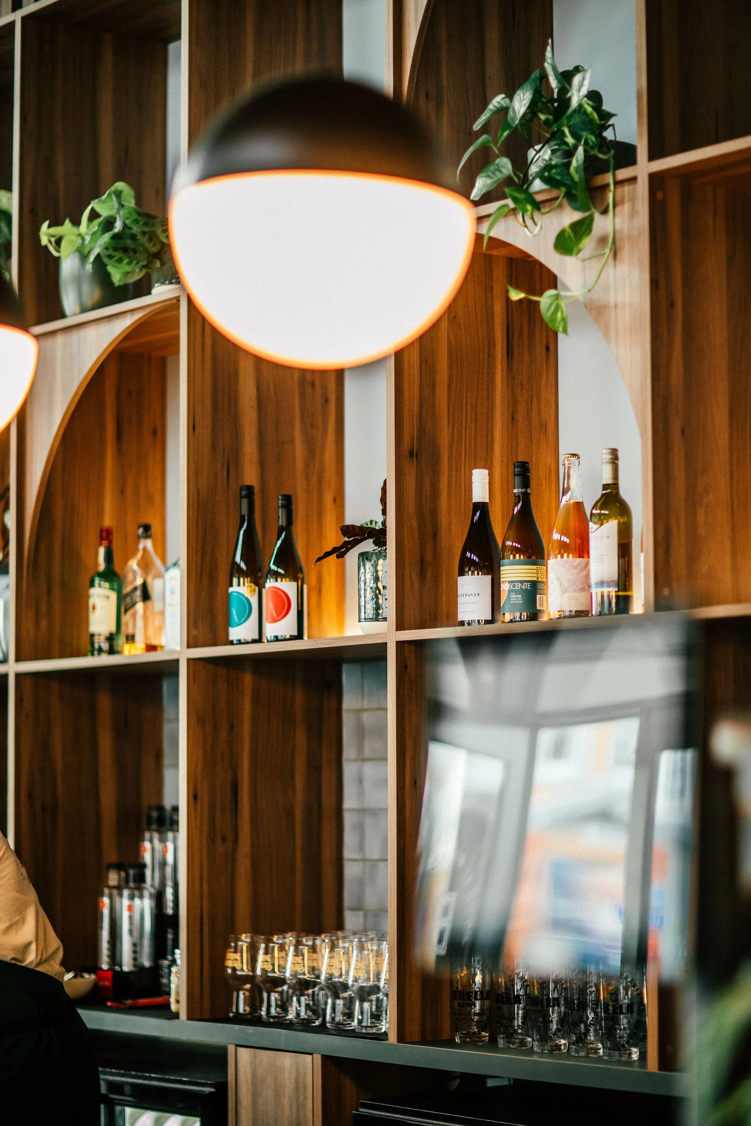 Wooden shelves holding wine bottles, plants, and glassware, illuminated by hanging orange-edged lights.
