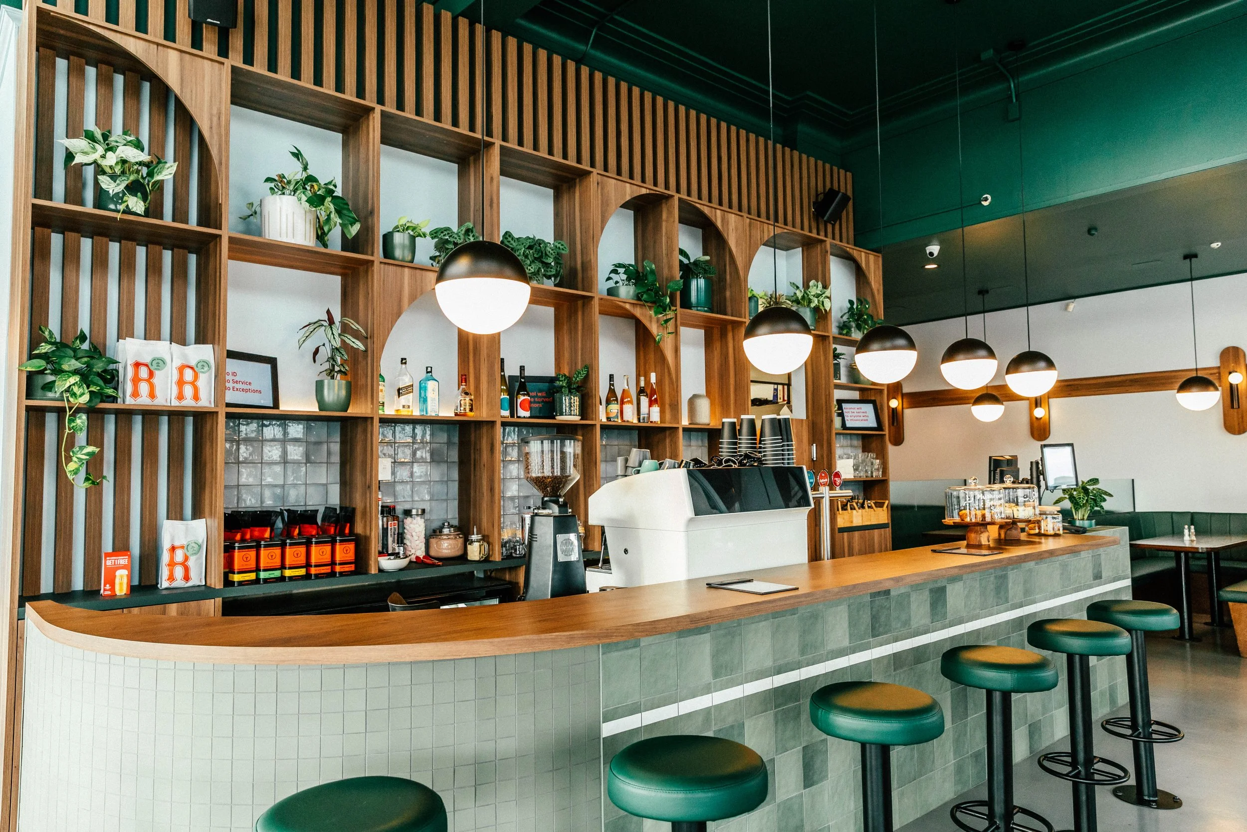 Interior of a modern café with a wooden counter, bar stools, a coffee machine, shelves with bottles and plants, and warm hanging lights.