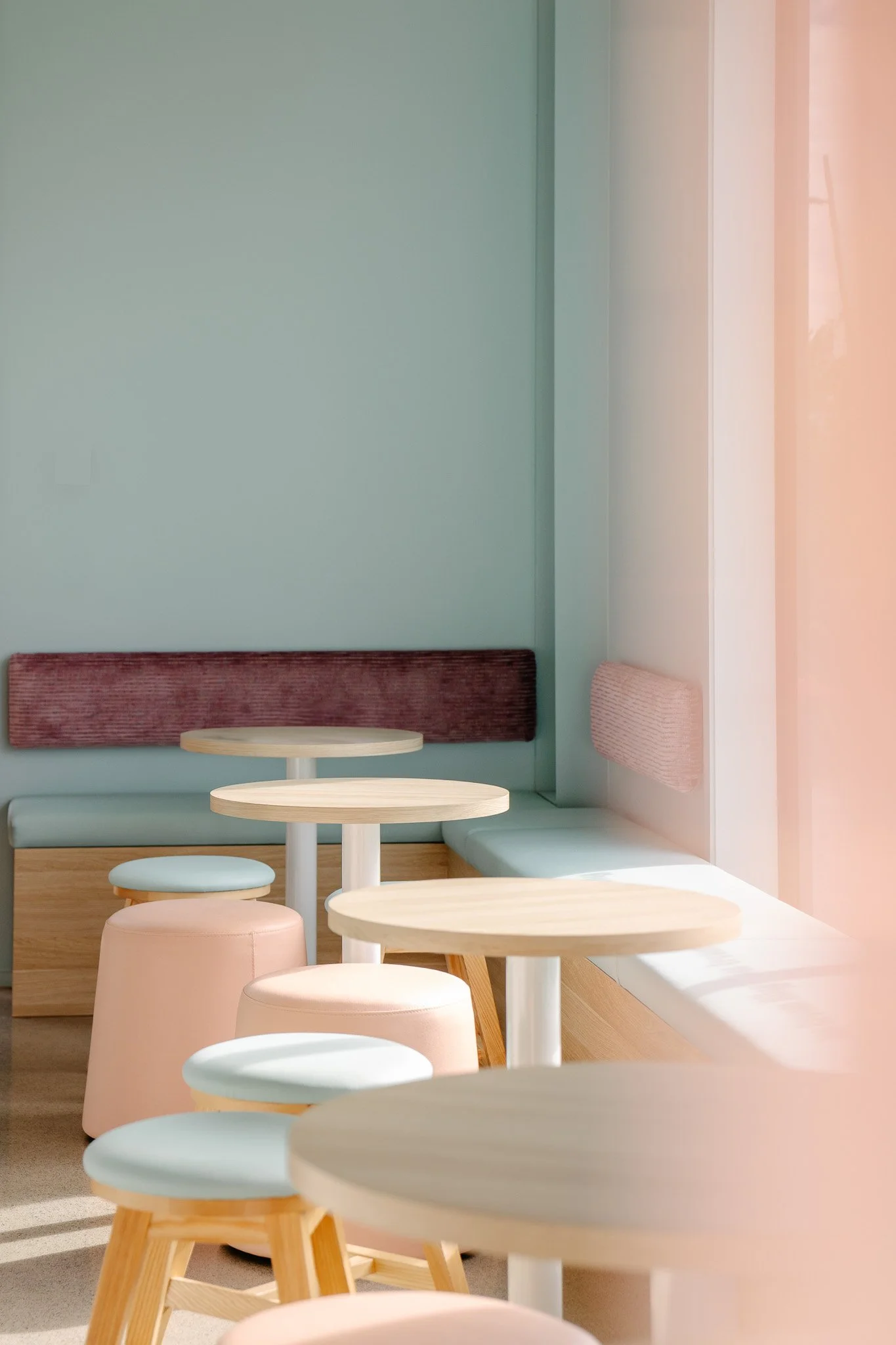 Interior of a coffee shop or café with pastel-colored furniture, light wood tables, round stools, and a cushioned bench seat near the window with sunlight streaming in.