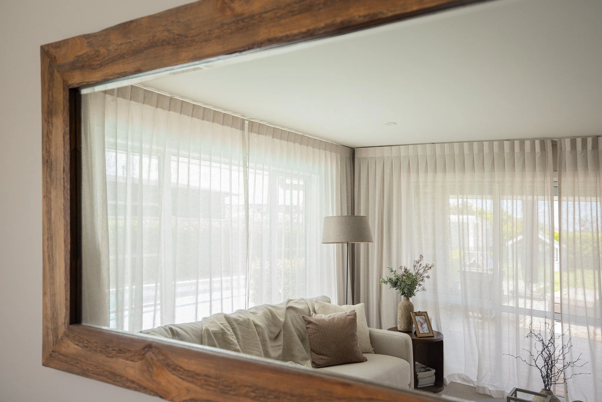 Living room seen through a large wooden-framed mirror, featuring a white sofa, beige cushion, floor lamp, vase with flowers, and sheer curtains allowing natural light.