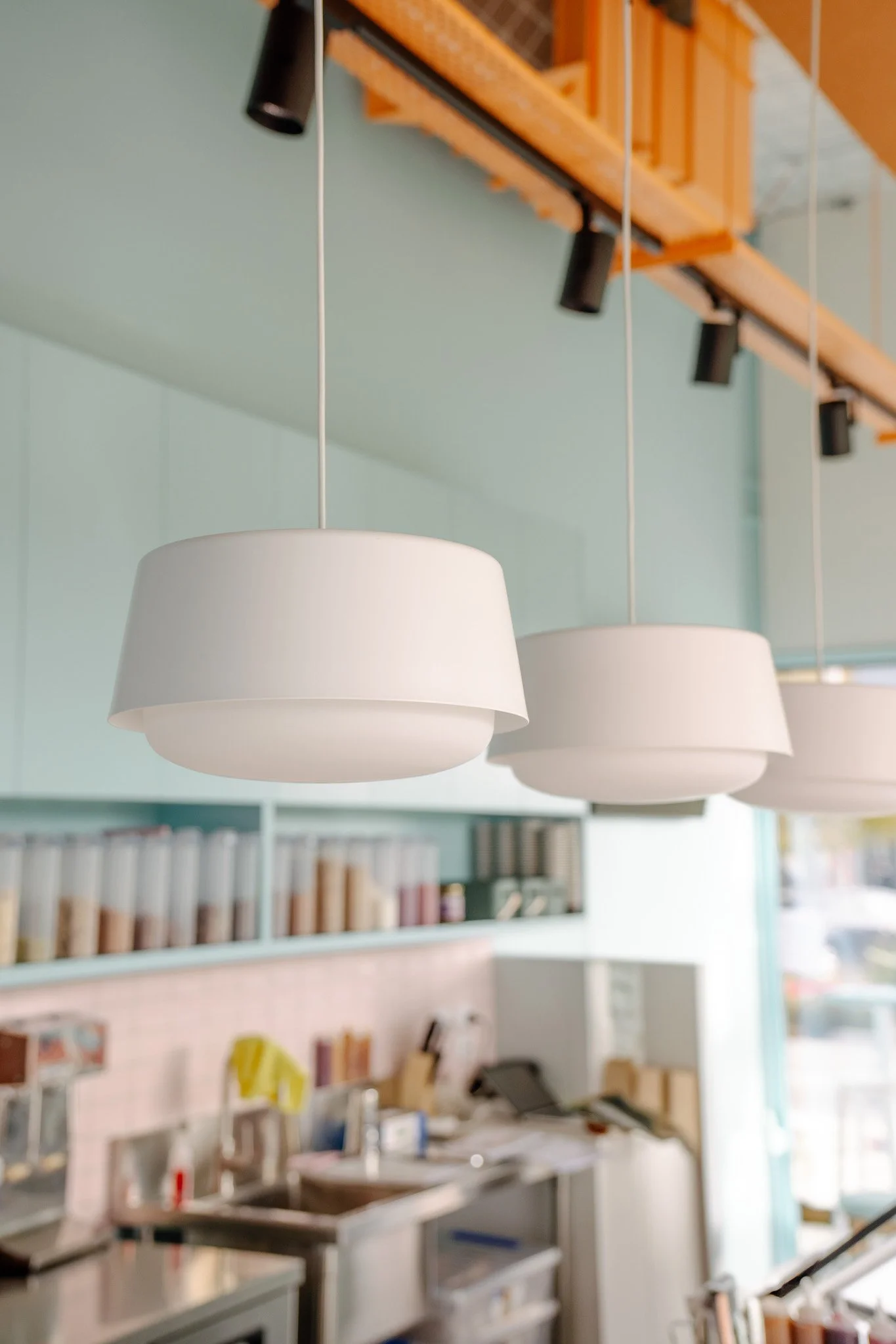 Three white pendant lights hanging from a ceiling above a kitchen or cafe counter with shelves of books or binders in the background.