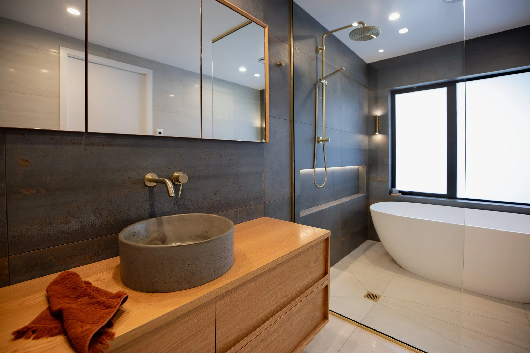 Modern bathroom featuring a freestanding bathtub by a large window, a walk-in shower with a rain showerhead, a concrete vessel sink on a wooden vanity, a large mirror, and dark tiled walls.