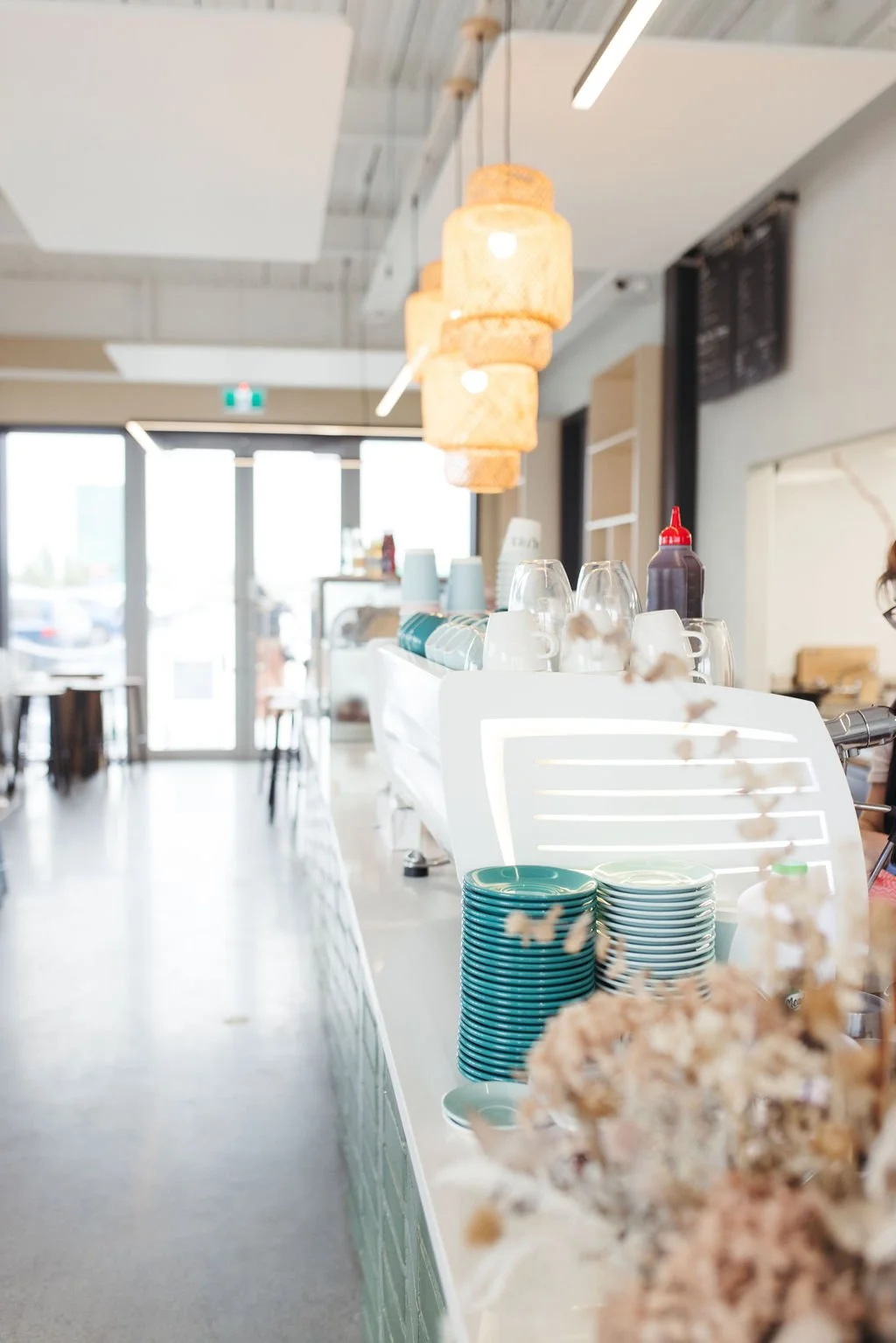 Interior of a bright, modern coffee shop with hanging woven lamps, stacks of teal and beige plates, cups, and a condiment squeeze bottle on the counter, and large windows letting in natural light.