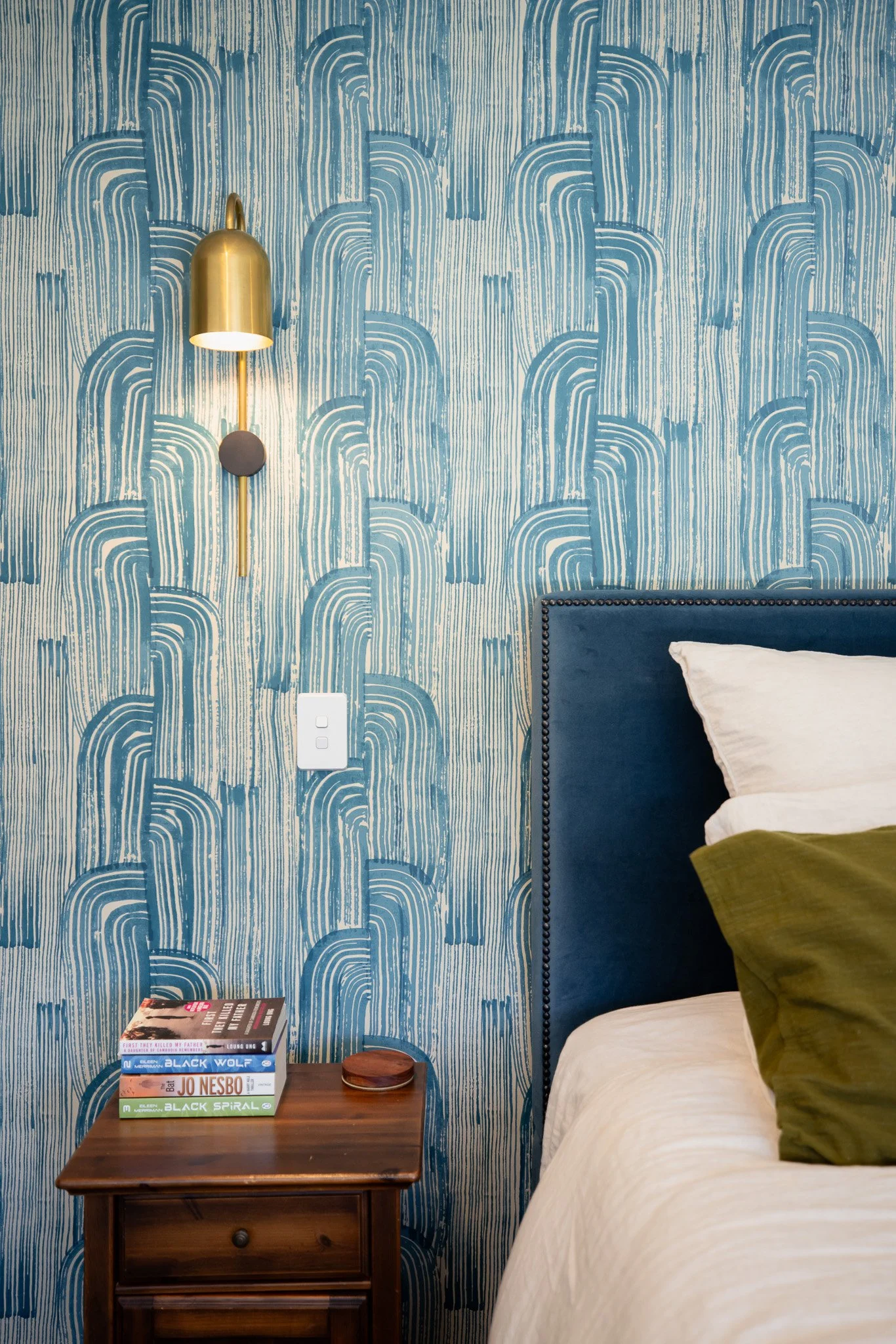 A bedroom corner with a blue upholstered bed, white pillow, green pillow, a wooden nightstand with three books, a decorative dish, a blue and white patterned wallpaper, and a gold wall sconce with a downward-facing light.
