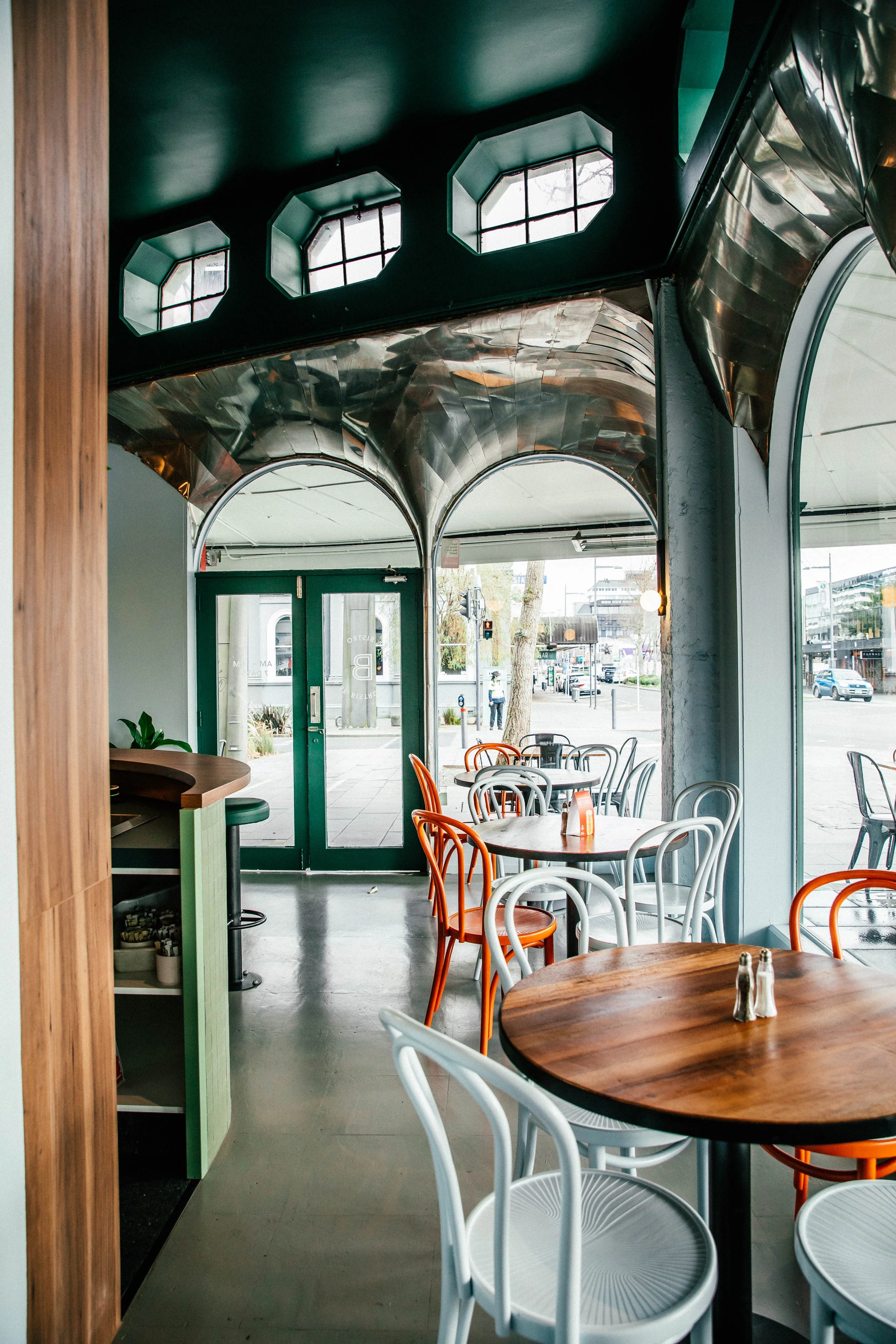 Interior of a modern restaurant or café with round wooden tables and colorful chairs, large windows, and a shiny metallic ceiling design.