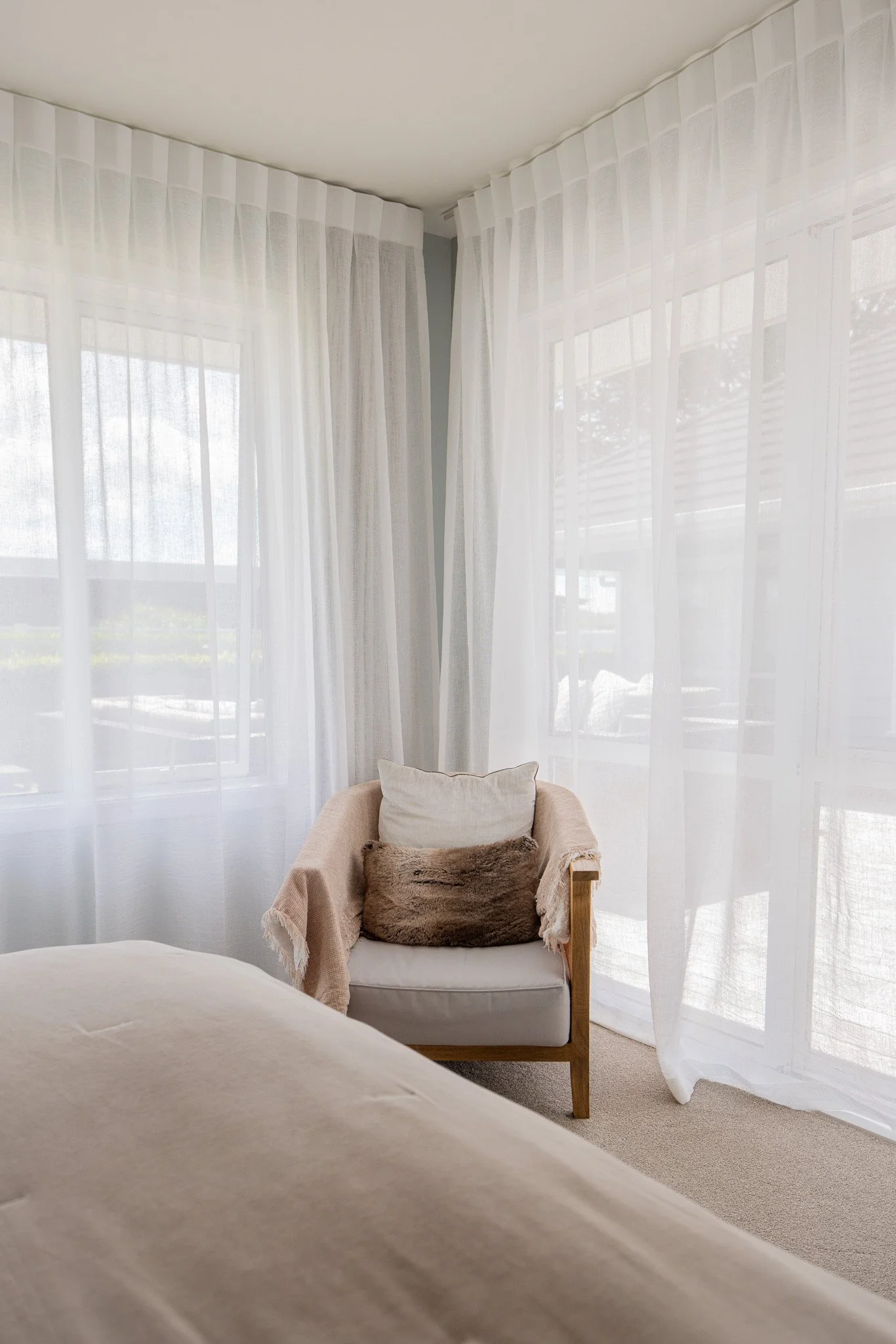 A cozy corner of a bedroom with a white armchair topped with a pillow, surrounded by sheer white curtains letting in natural sunlight.