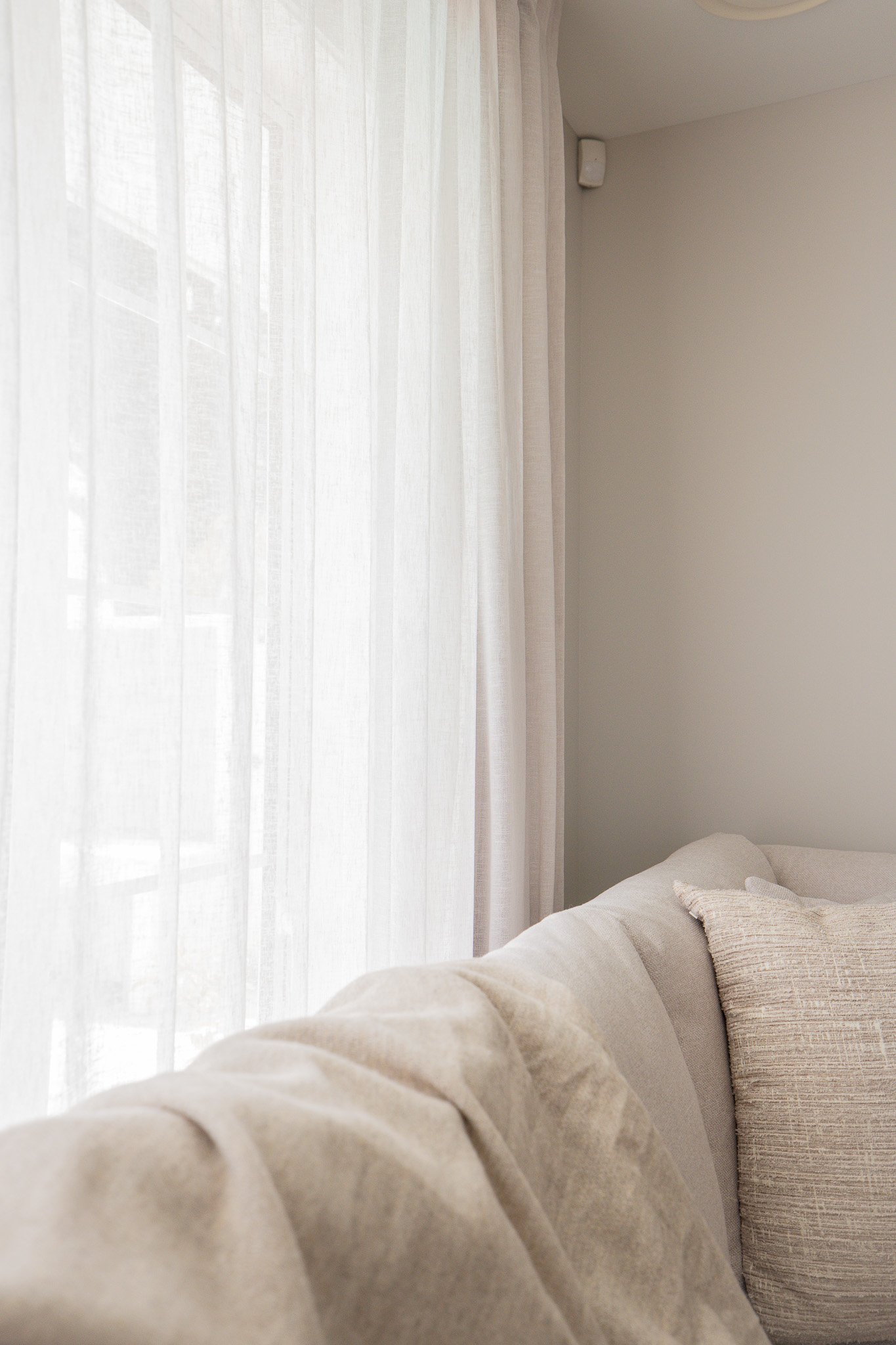 A living room with beige couches, a throw pillow, and sheer white curtains covering a window.