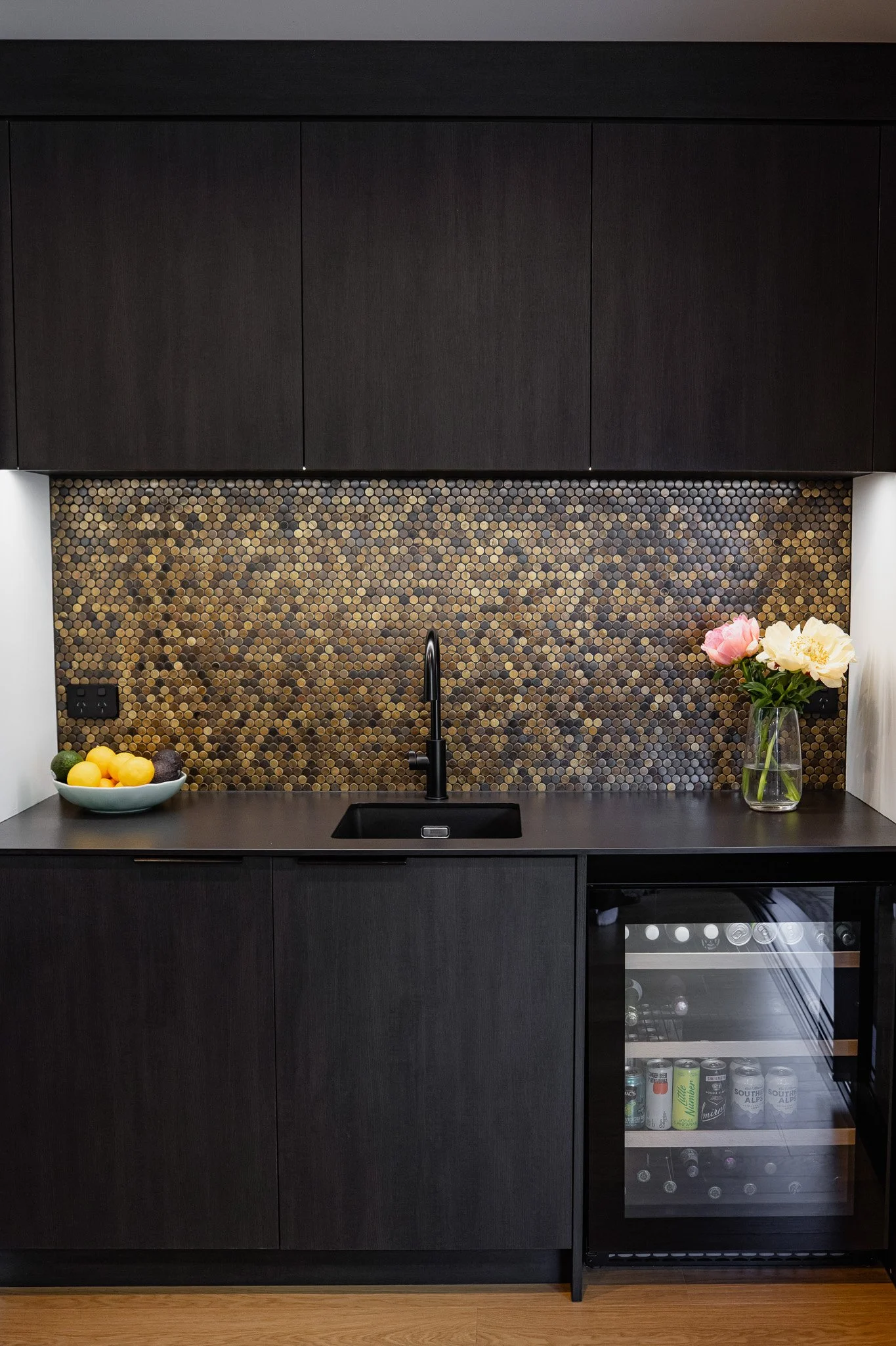 Modern kitchen with black cabinetry, a black backsplash with small round tiles, a black sink with a black faucet, a bowl of lemons and limes, and a vase with pink and white flowers.