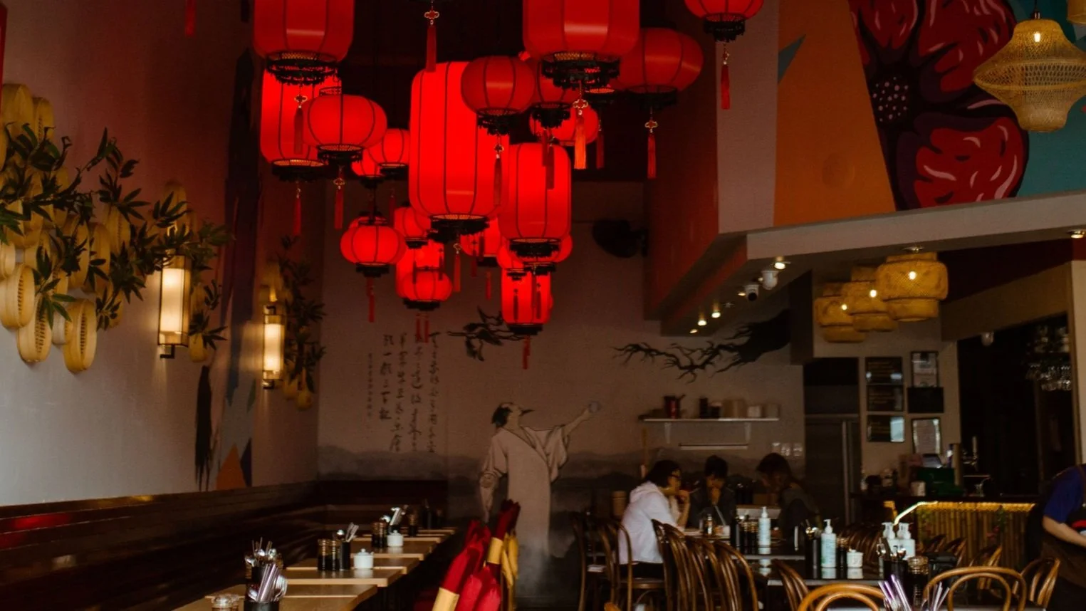 Inside a restaurant with red lanterns hanging from the ceiling, wall art featuring a woman in traditional attire, and diners seated at tables.