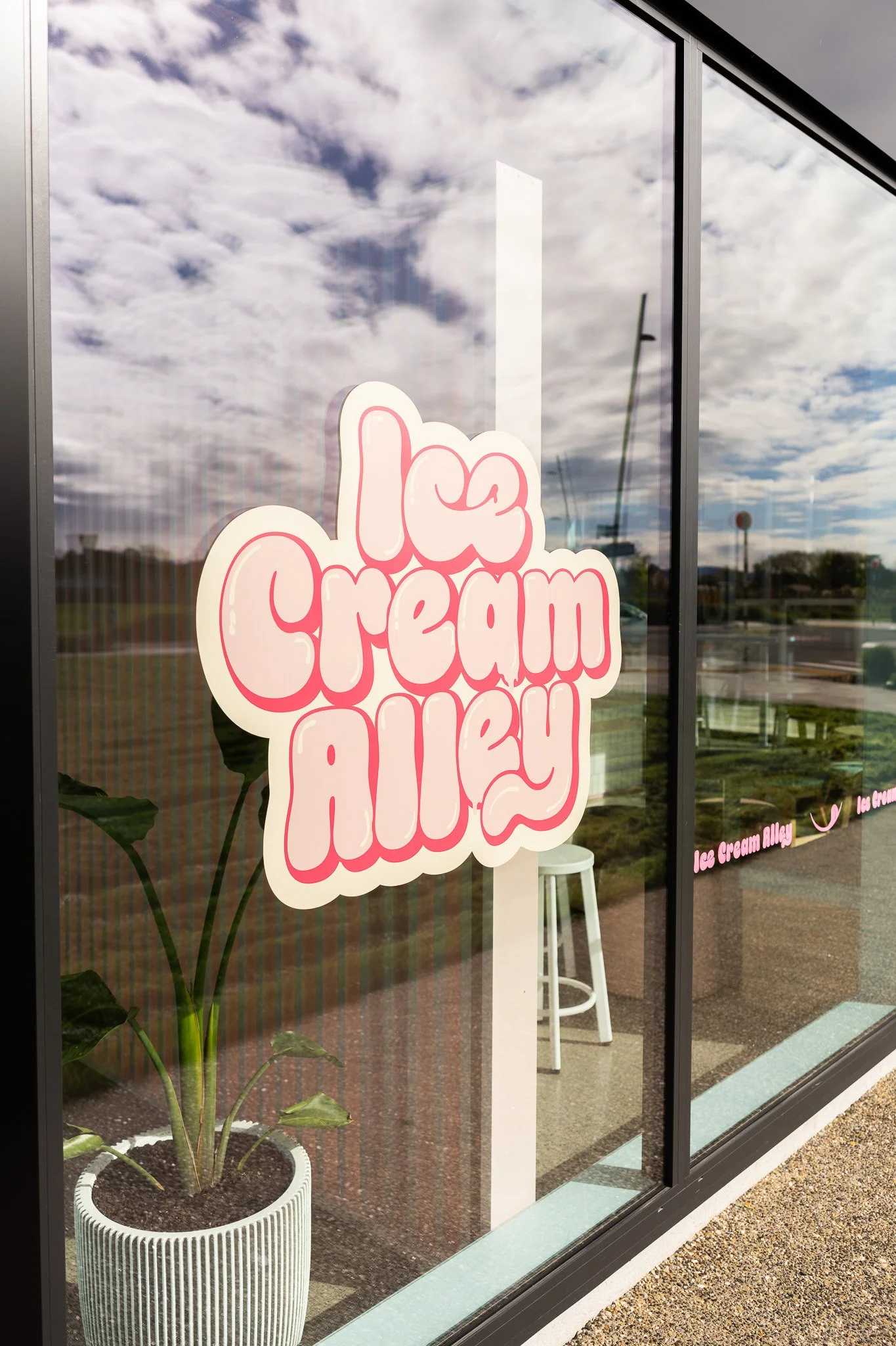 Storefront window with pink and white sign reading "Ice Cream Alley," a white stool, and a potted plant with large green leaves outside, reflecting a partly cloudy sky.