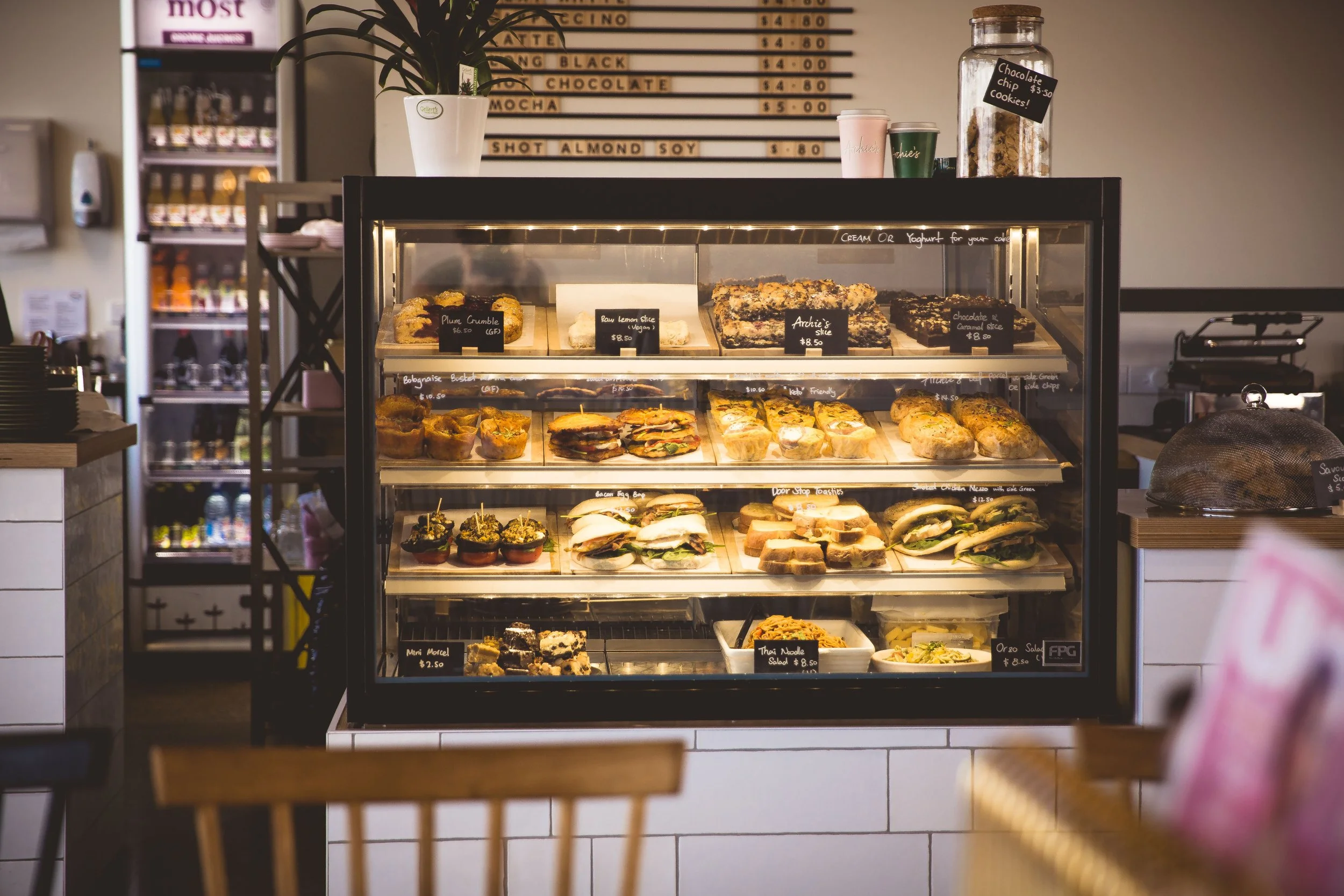 Display case filled with assorted sandwiches, baked goods, and desserts in a cafe. A menu board hangs behind with prices, and there are cups and containers on top of the display case.