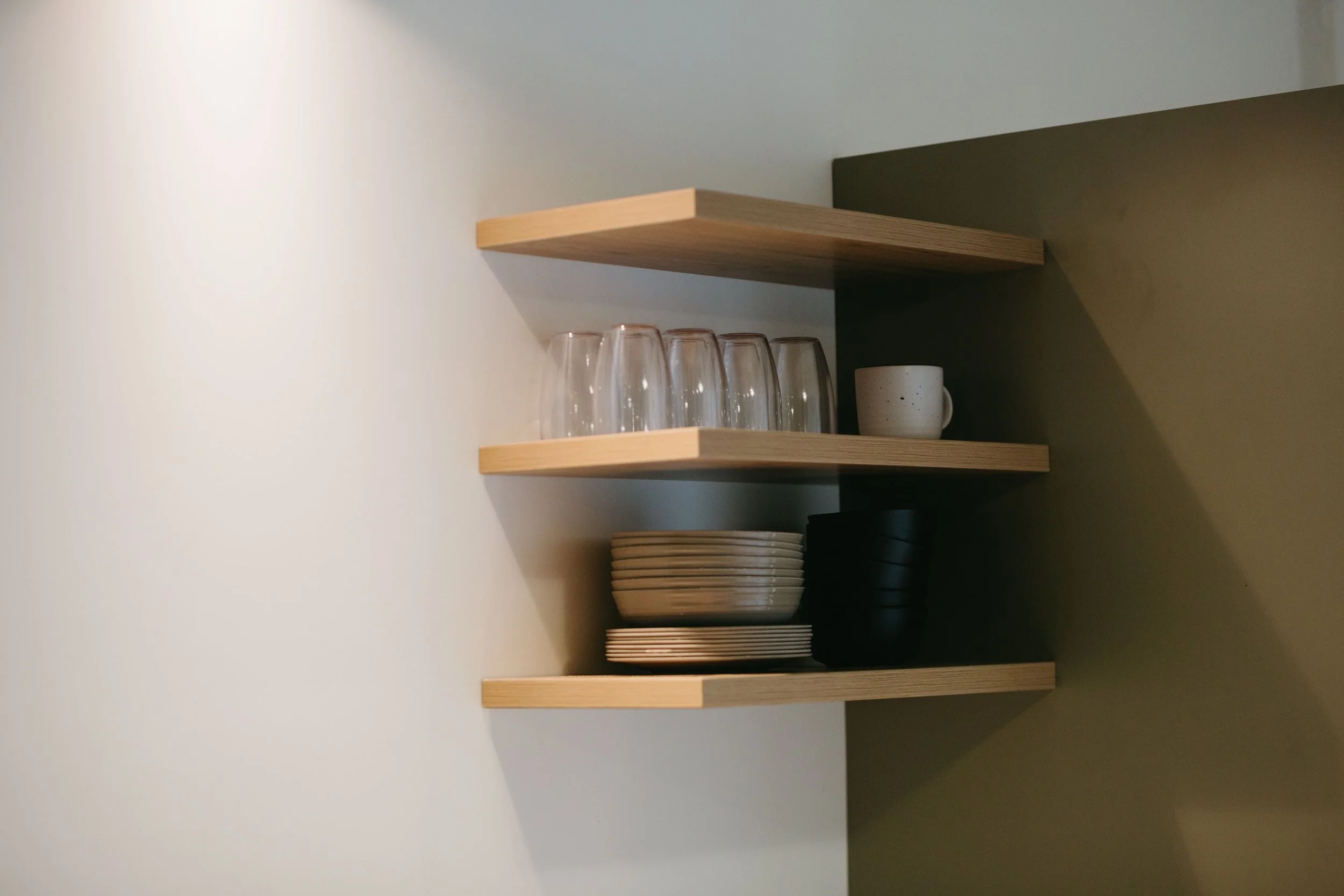 Wall-mounted wooden shelves holding glassware, plates, and bowls in a minimalist kitchen.