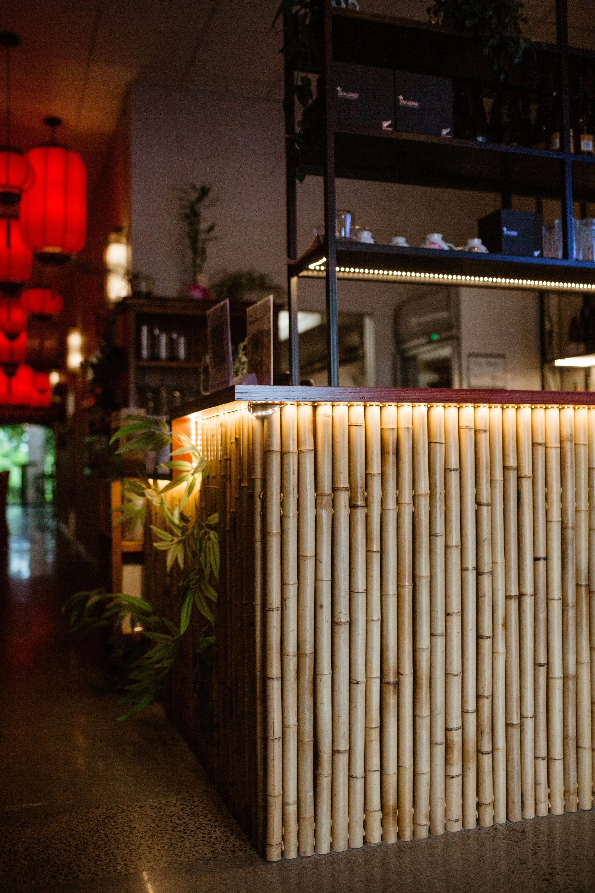 Interior of a restaurant or cafe with a bamboo-covered counter, red lanterns hanging from the ceiling, and plants near the reception area.