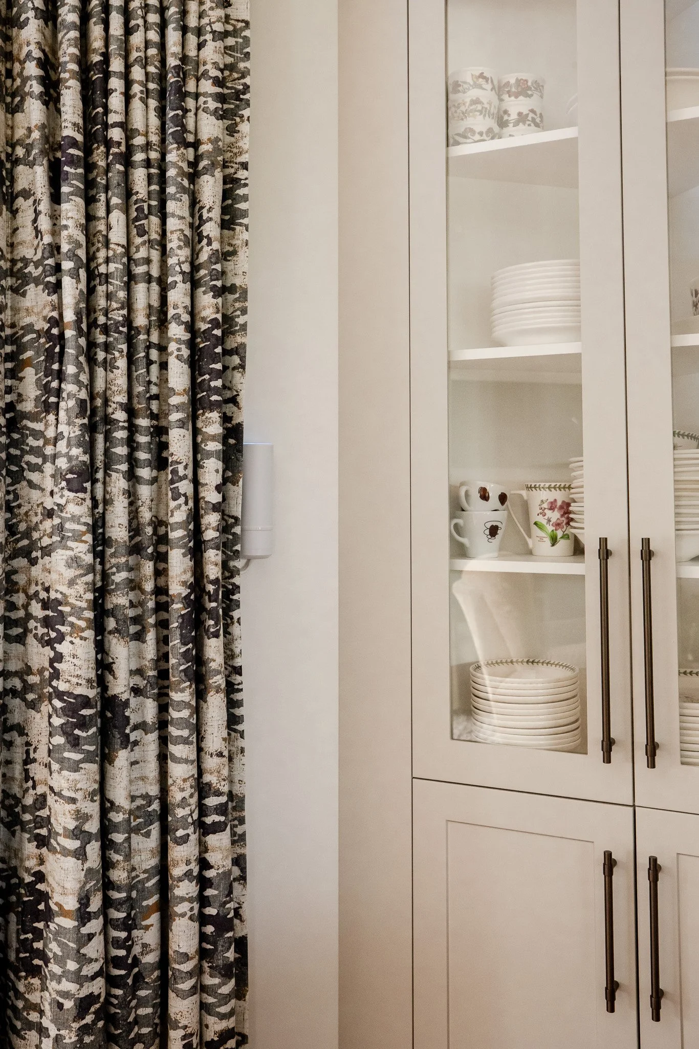 A kitchen cabinet with glass doors displaying white plates, bowls, and cups, next to a patterned curtain.