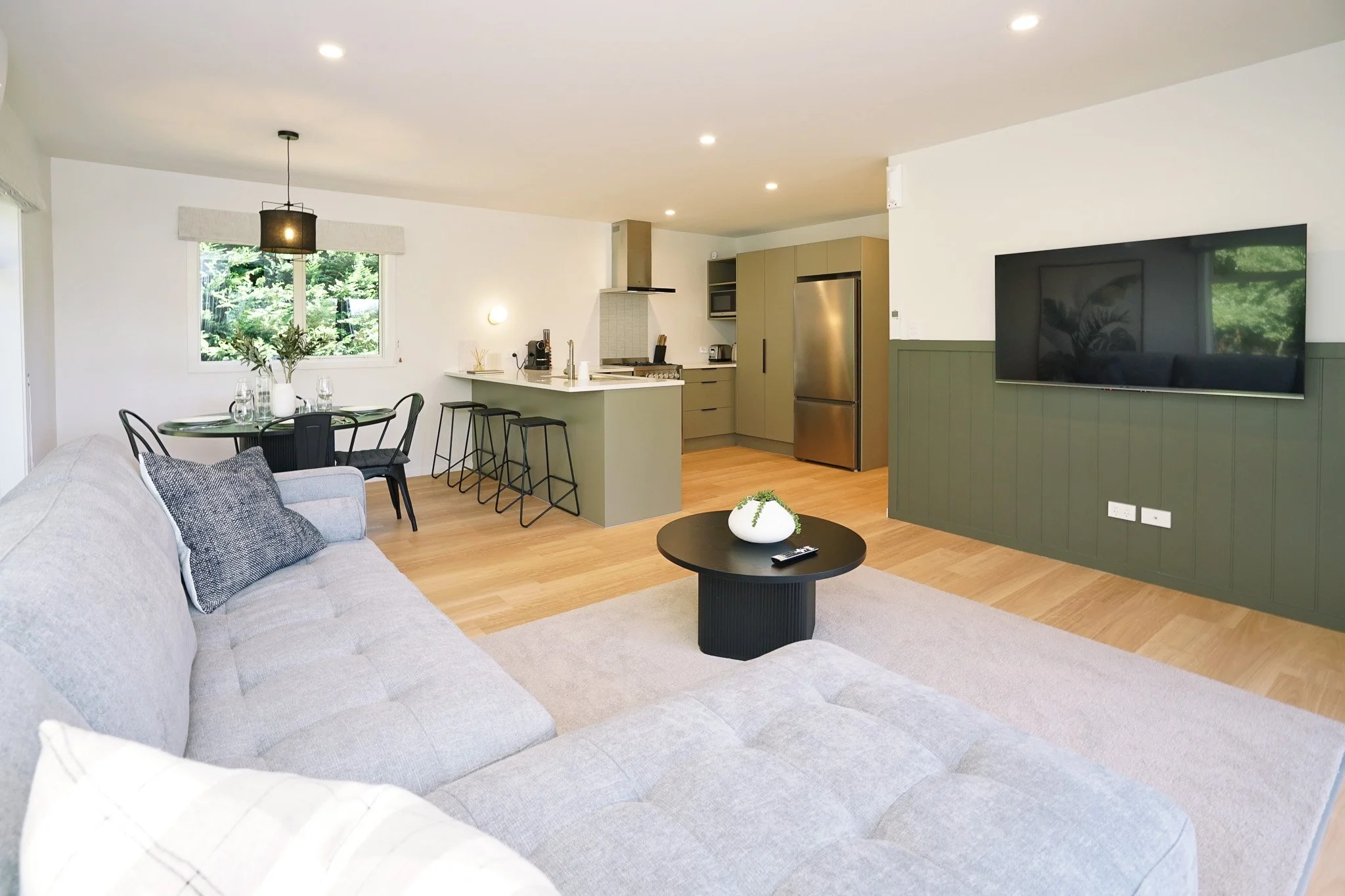 Living room with light gray sectional sofa, black coffee table with white vase, TV mounted on green paneled wall, open kitchen with beige cabinetry and stainless steel appliances, dining table with six black chairs, large window with outside greenery.