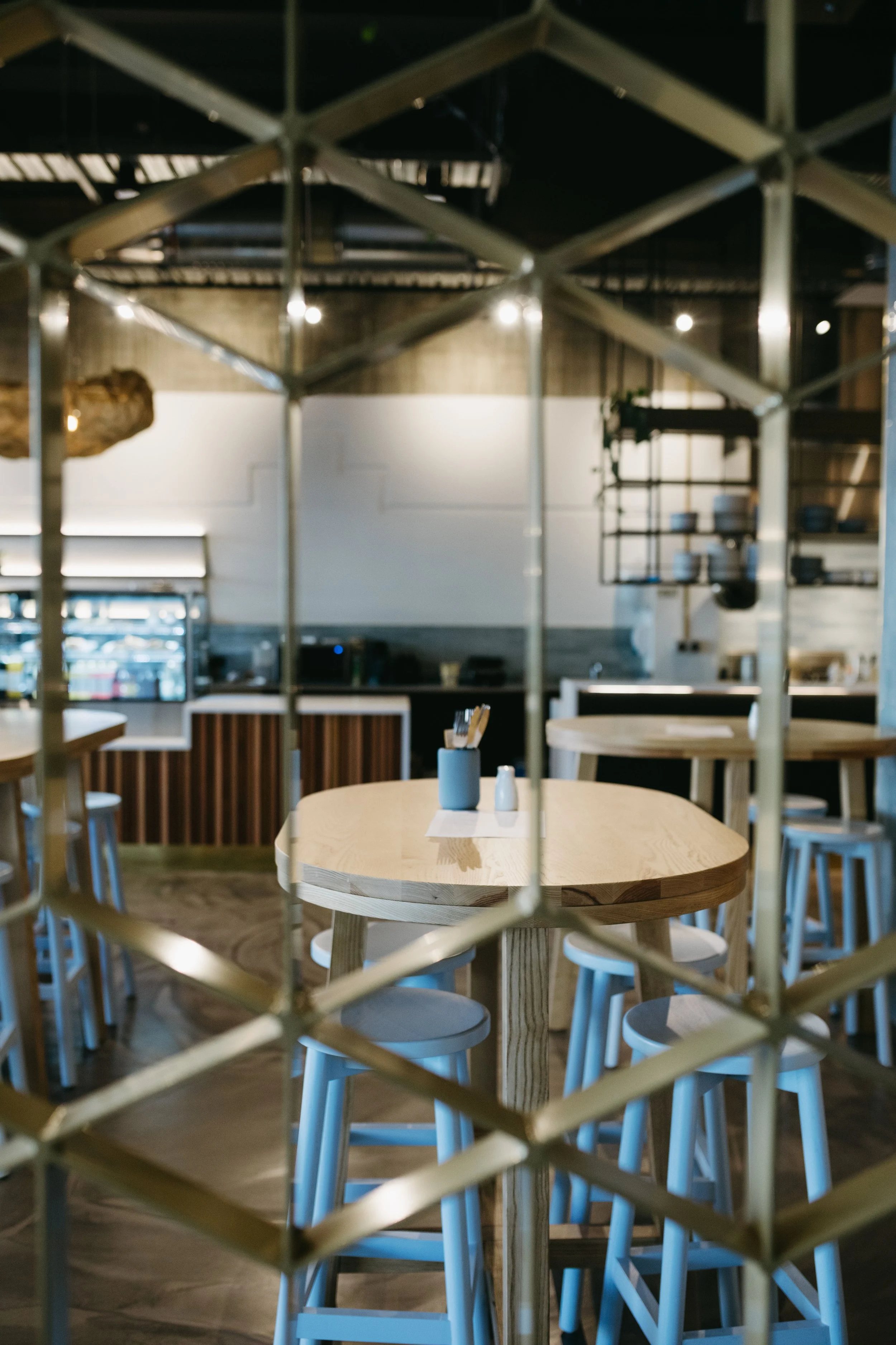 View of a modern, minimalist restaurant interior through a lattice partition, featuring a round wooden table with stools, and a background with a counter, shelves, and ambient lighting.