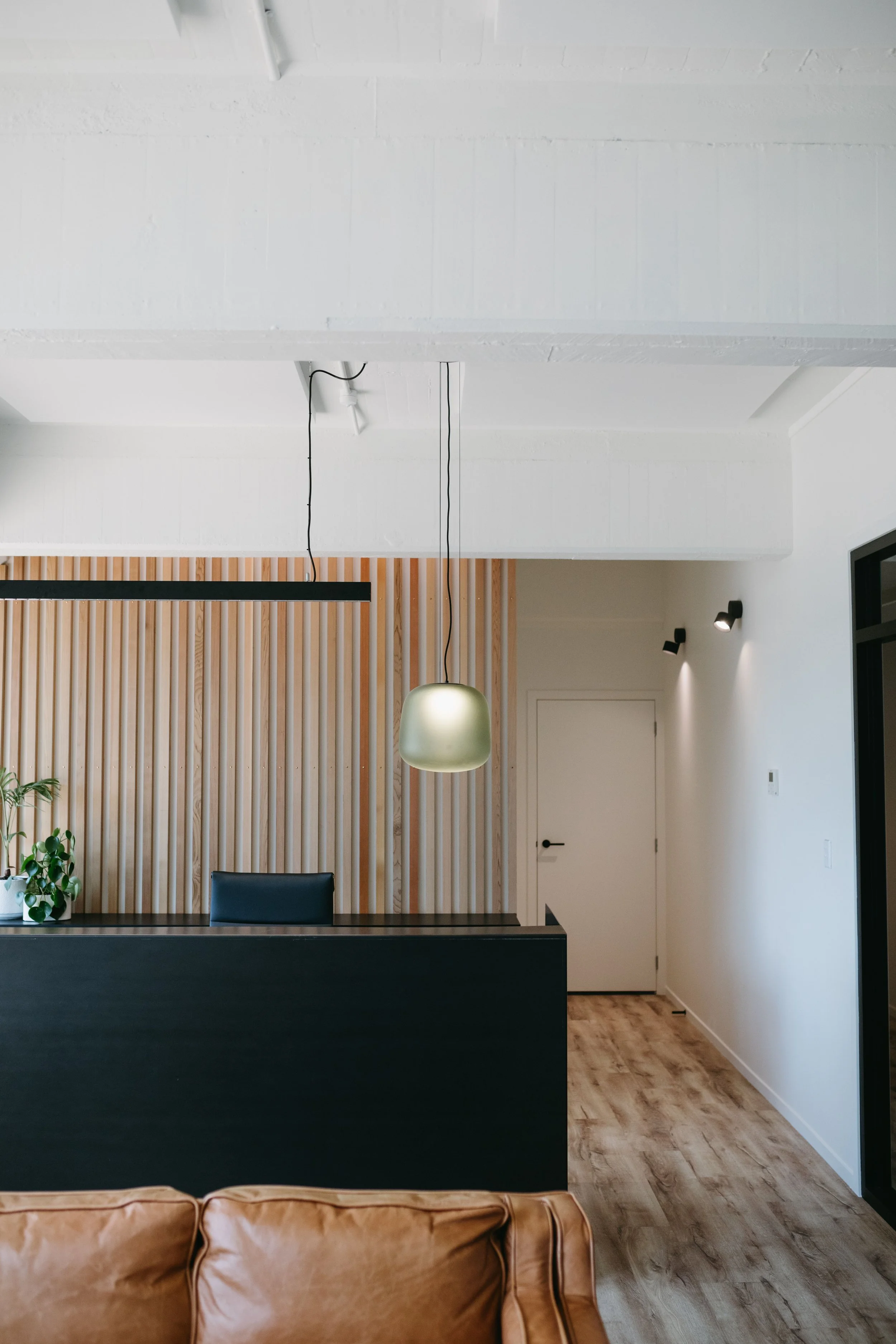 Modern interior with wooden wall paneling, black reception desk, hanging pendant light, leather couch, and white walls.
