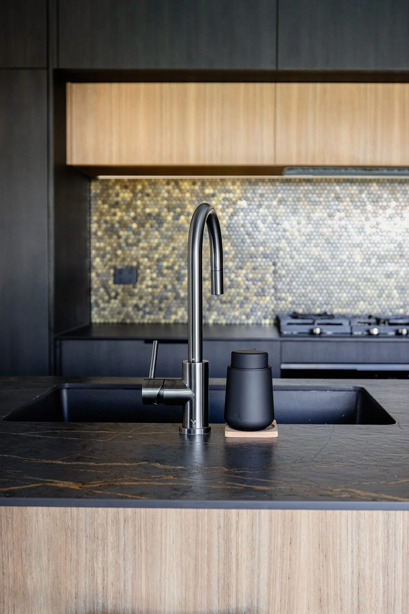 Modern kitchen with black sink, chrome faucet, black soap dispenser, and a mosaic tile backsplash with gold, black, and gray tiles.
