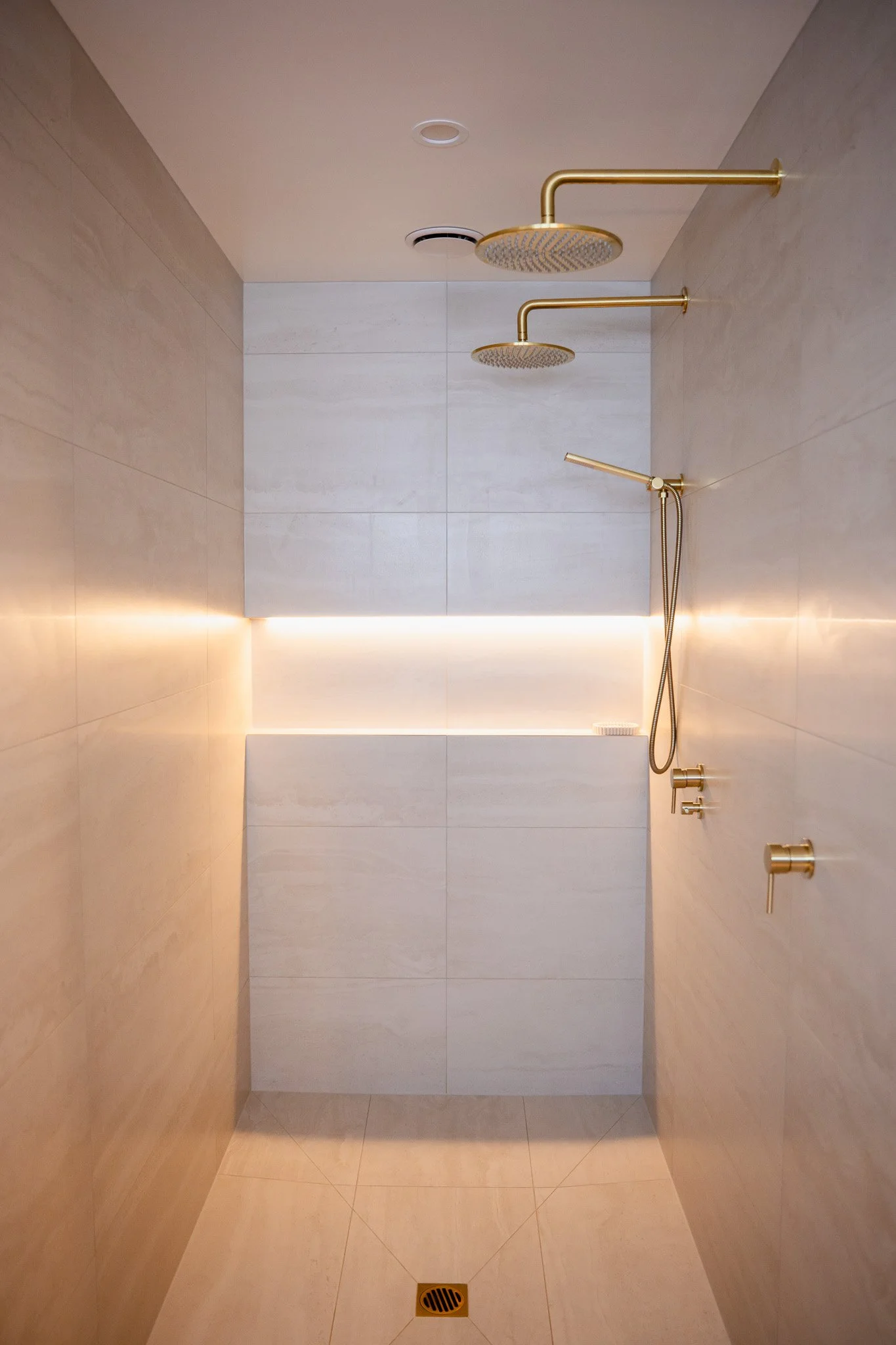 Modern shower with two overhead rain shower heads, a handheld shower head, and sleek brass fittings and controls in a minimalist design bathroom with light-colored tiles and ambient lighting.
