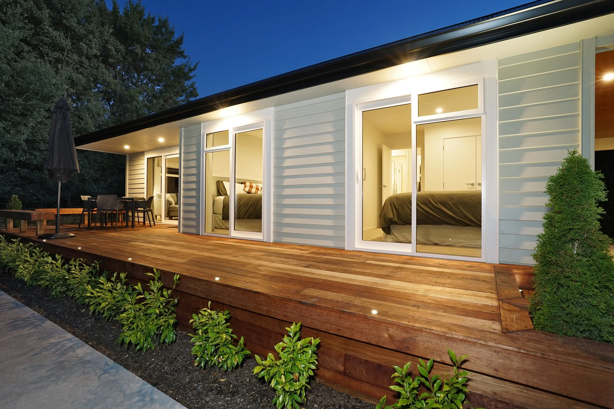 Modern house with large glass sliding doors opening onto a wooden deck, outdoor furniture, plants, and trees visible in the background at dusk.