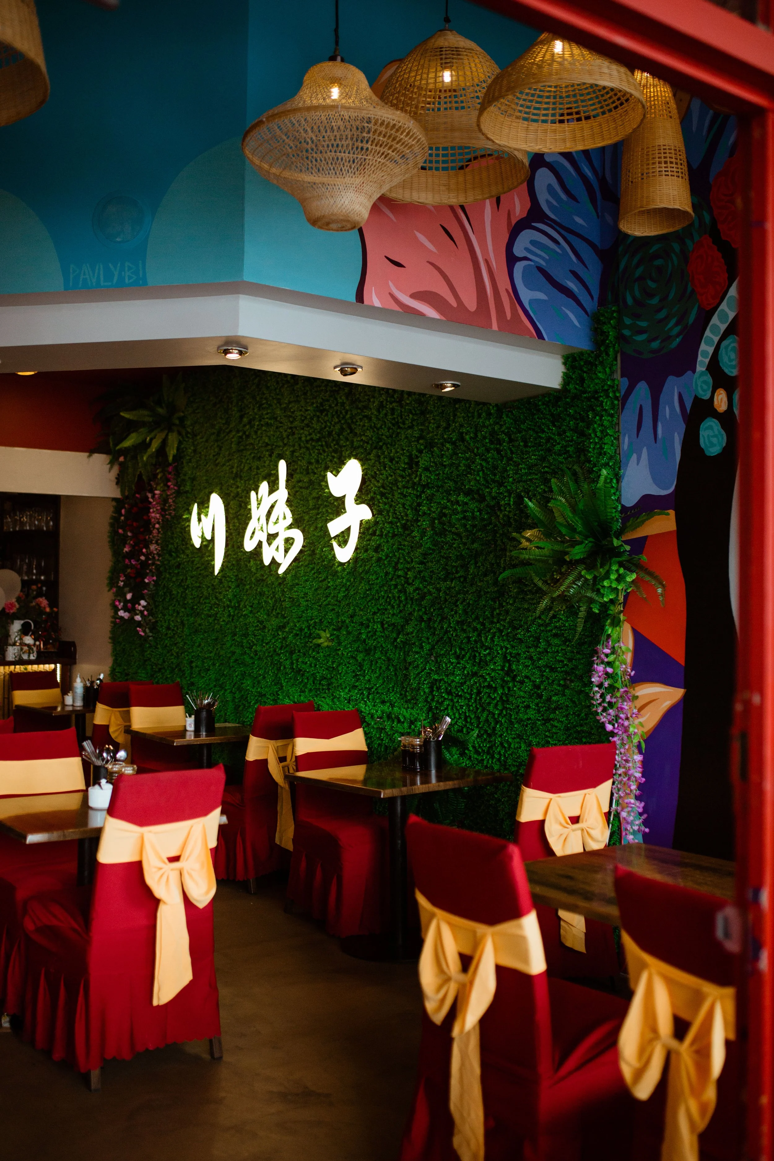 Interior of a restaurant with red chairs decorated with beige bows, a green wall with Chinese characters lit up, tropical plants, colorful murals, and woven pendant lights.