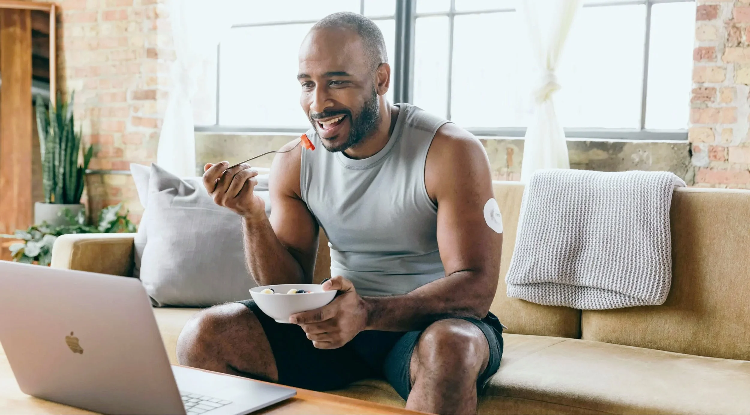 Fit man sits on couch, eating fruit as he takes part in a conversation via his laptop.